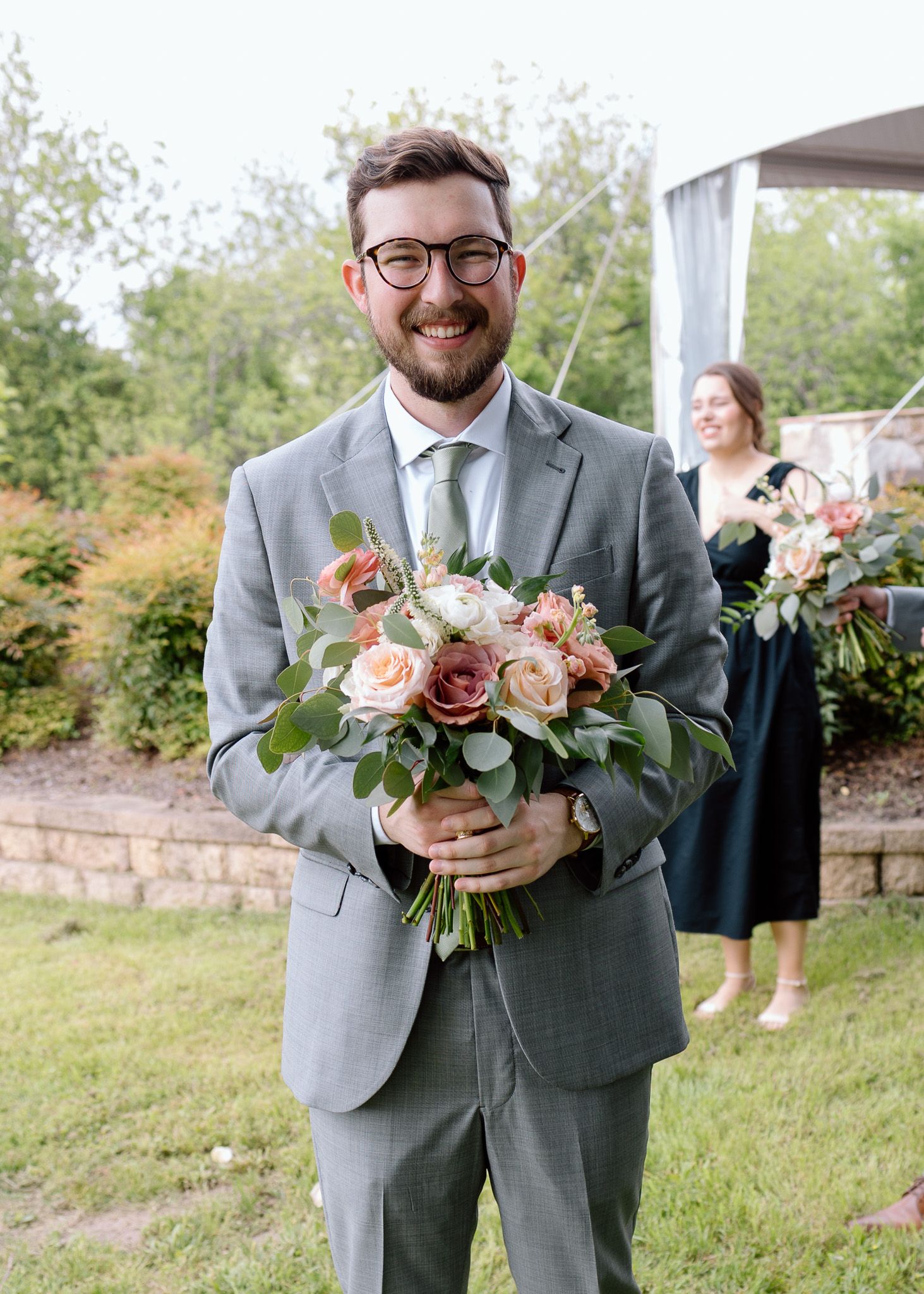 Wedding attendant in gray suit holds a beautiful bouquet of pink and white roses with greenery.