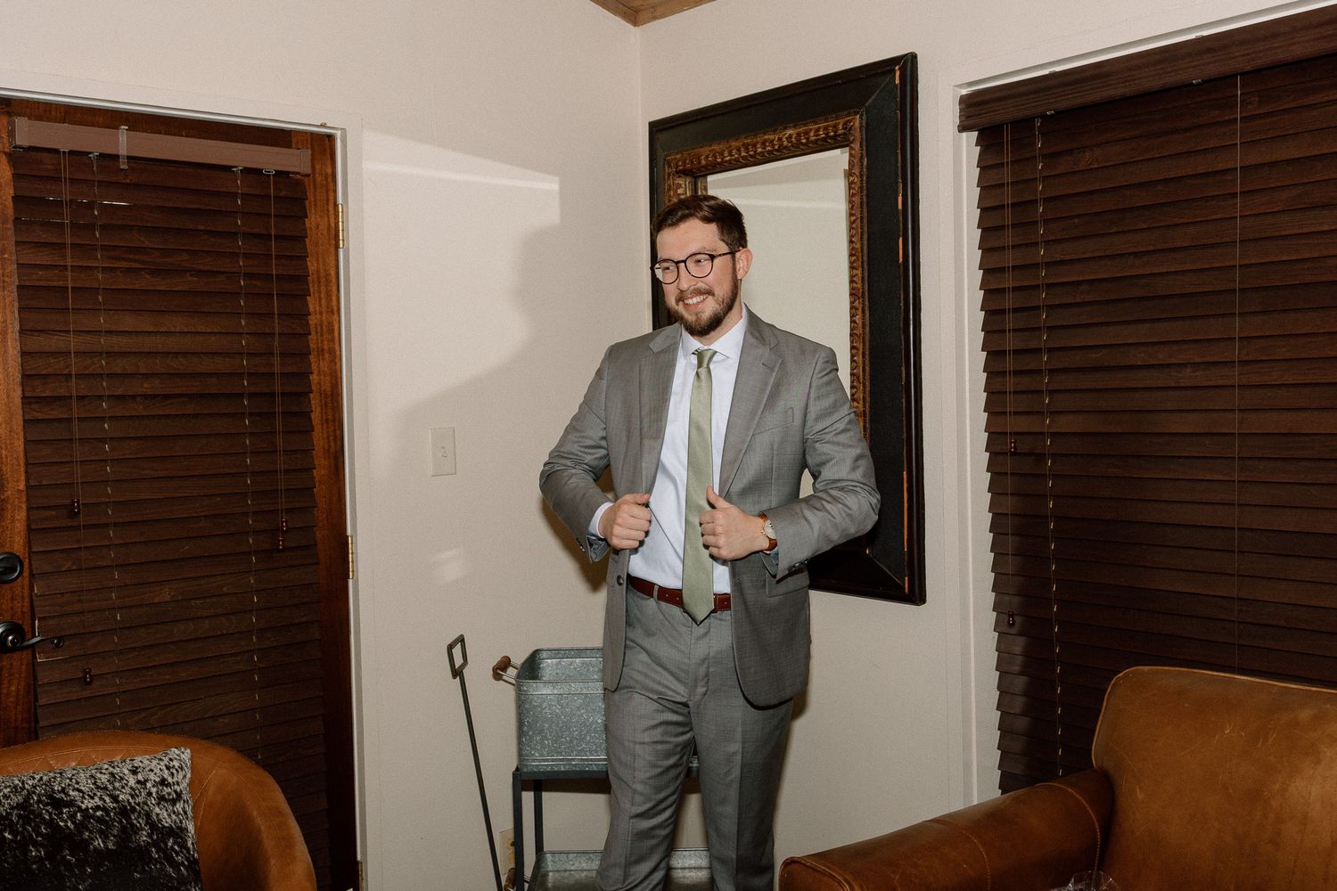 A person in a grey suit standing in a room with wooden window blinds and brown leather chairs.