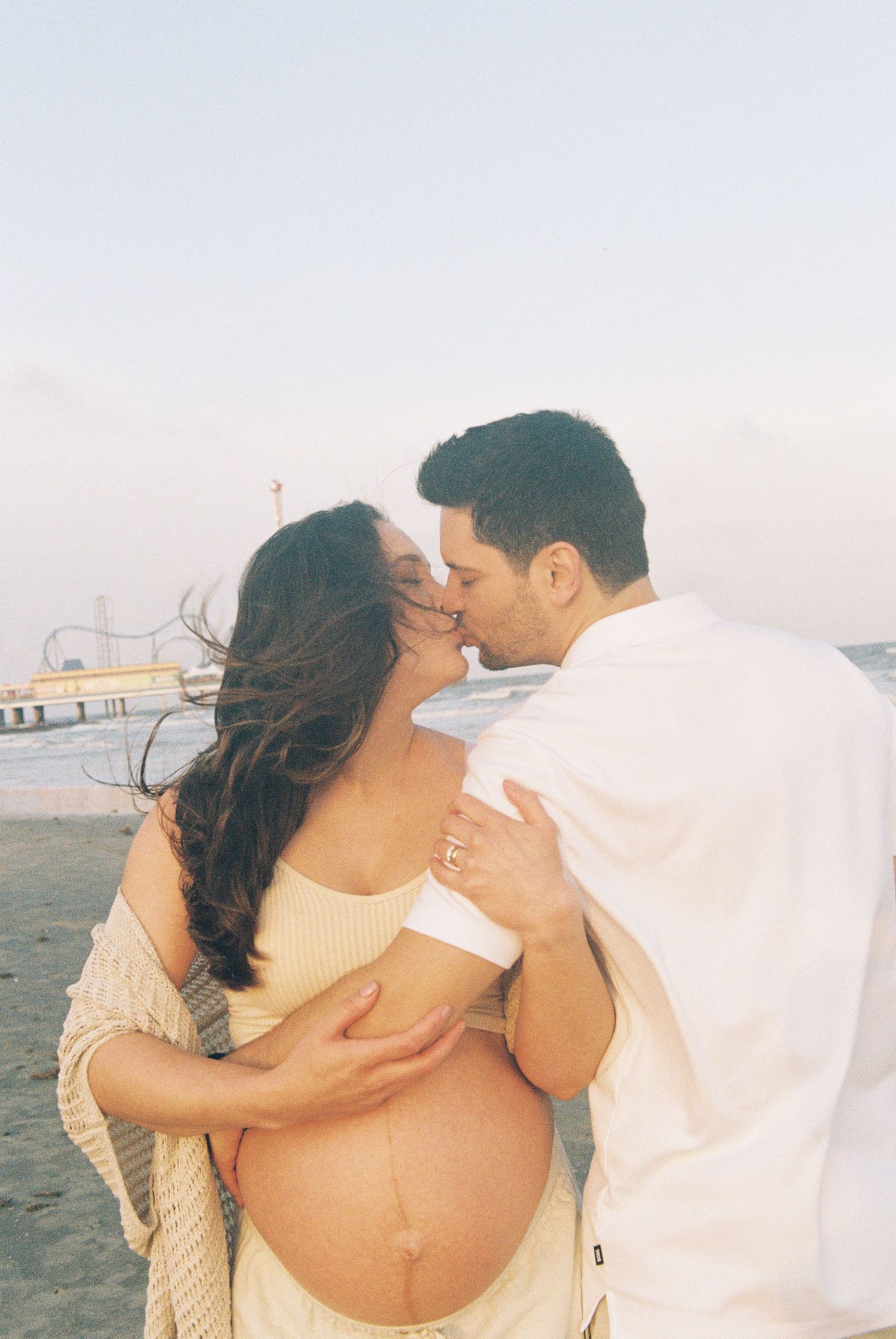 Romantic maternity photoshoot on the beach with a couple embracing at sunset with an amusement park pier in background.