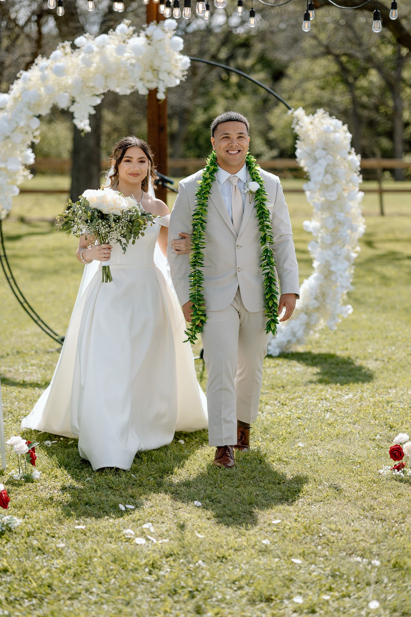 Newlyweds stand under a white floral arch wearing traditional wedding attire and green lei.