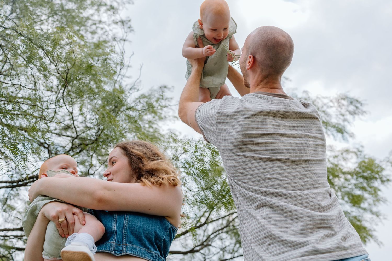 Family enjoying a playful outdoor moment together with their baby against a cloudy sky and tree backdrop.