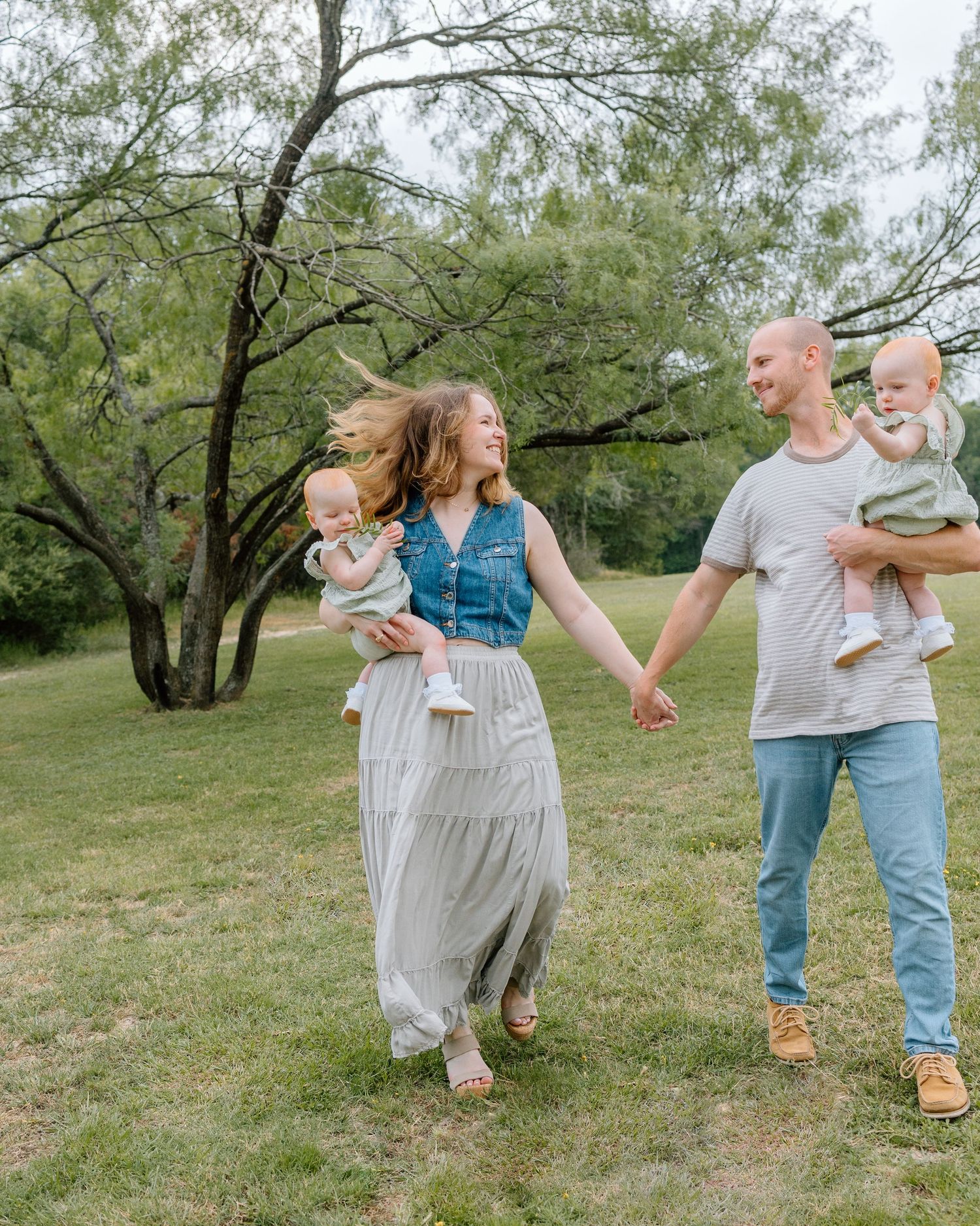 Happy couple walking in a spring garden while holding hands and carrying toddlers amid blooming trees.