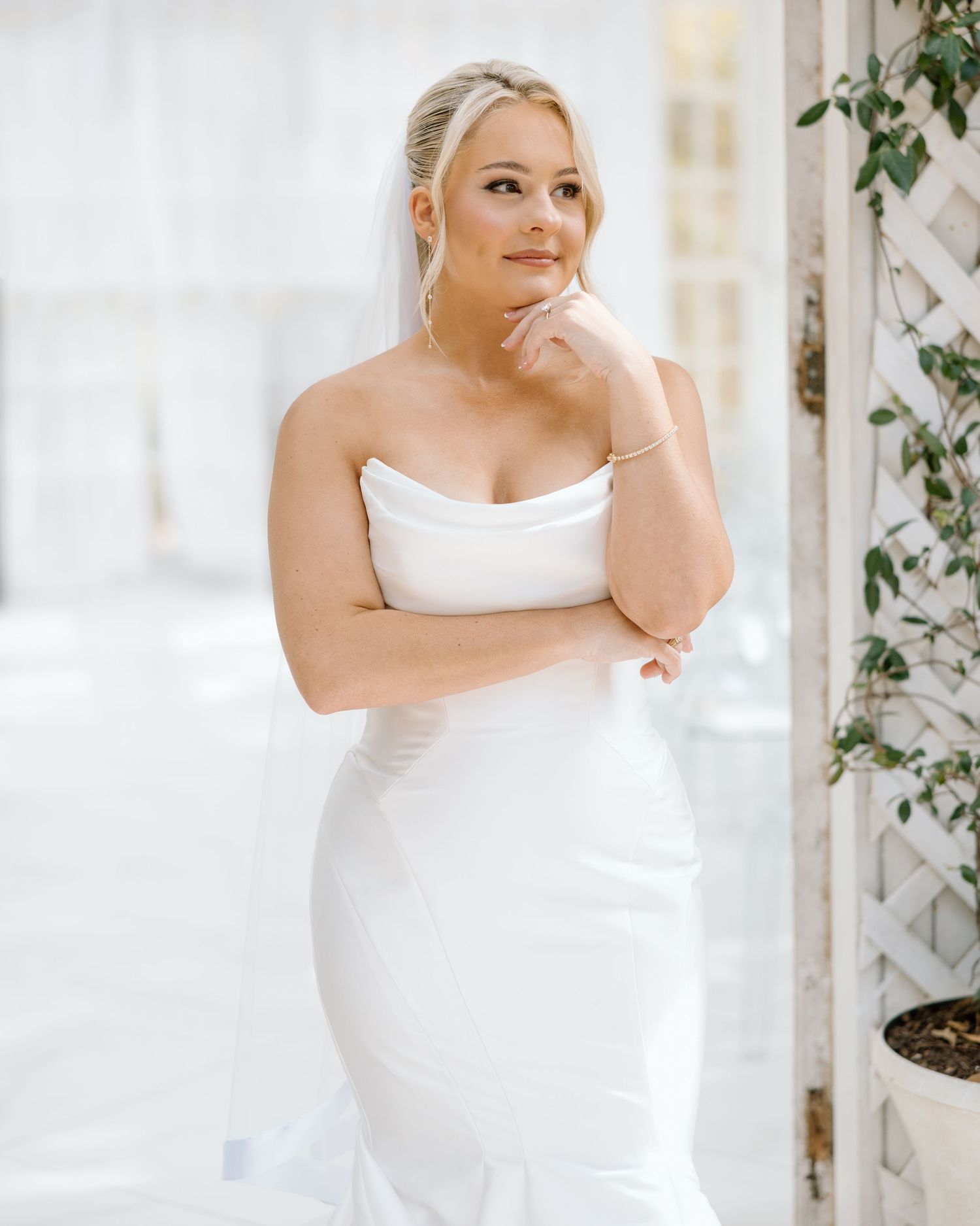 Elegant bride in strapless white mermaid wedding dress with veil poses thoughtfully in bright venue.