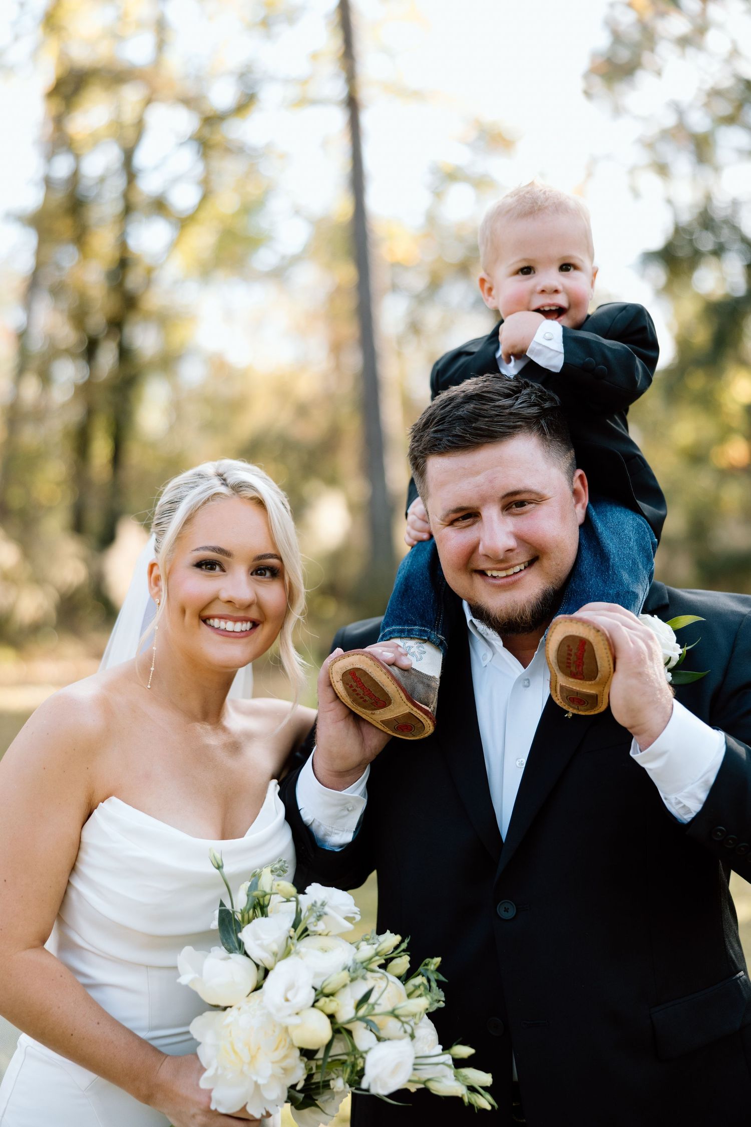 Happy newlyweds smiling with a baby perched on grooms shoulders during outdoor wedding portraits.