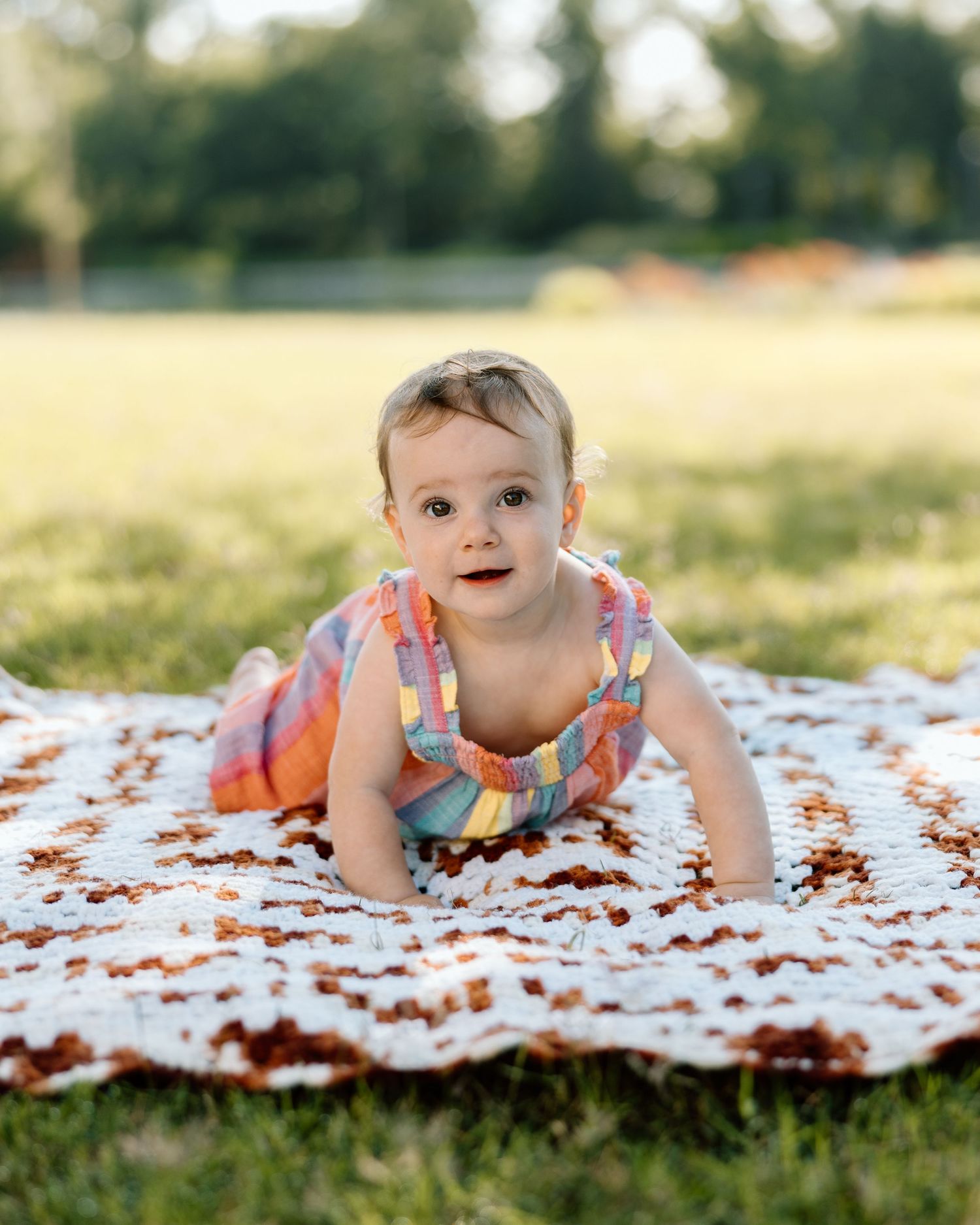 Baby girl in colorful dress doing tummy time on crochet blanket outdoors in green park.