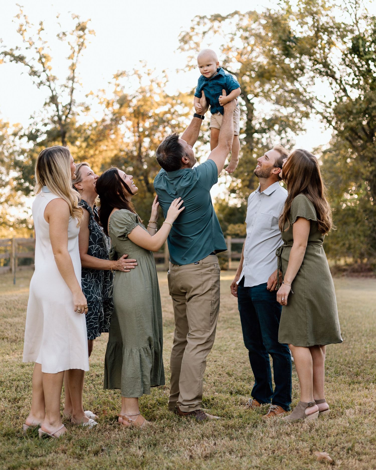 Family gathering outdoors lifting a baby in the air, smiling together on a grassy lawn with trees in the background.