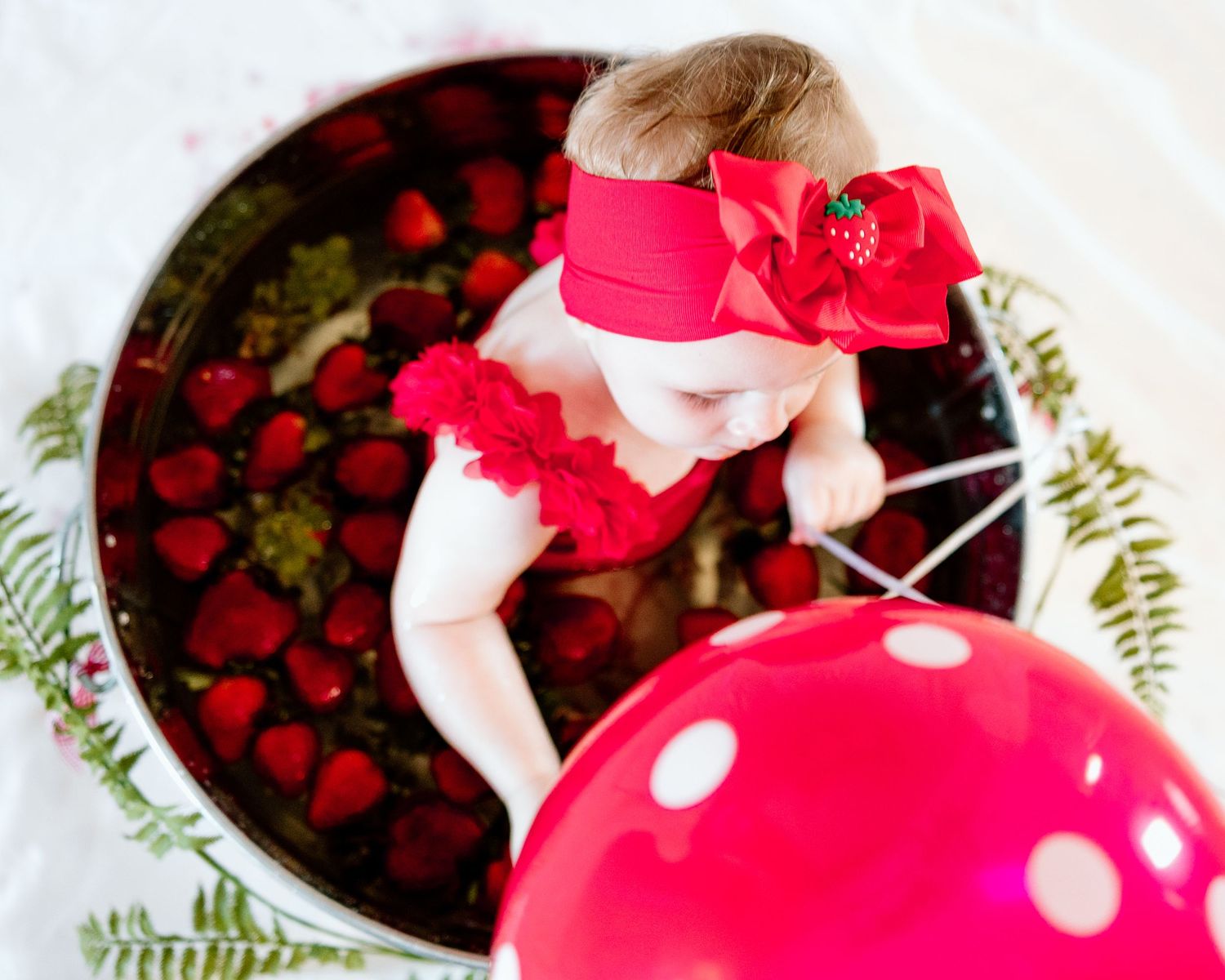 Baby girl in red ruffle outfit and bow headband sits in a tin filled with fresh strawberries near a red balloon.