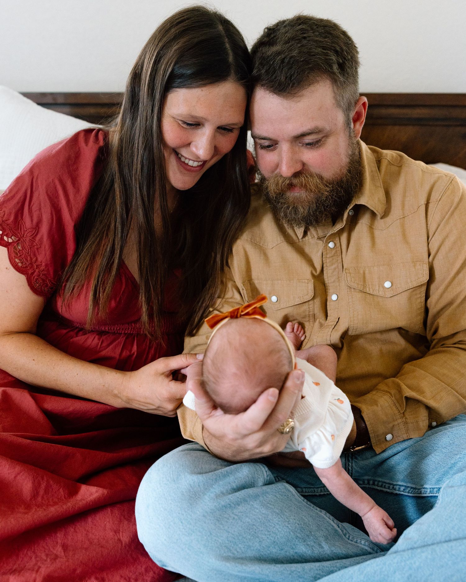 Smiling parents in red and tan outfits lovingly gaze at their newborn baby girl with an orange bow headband.