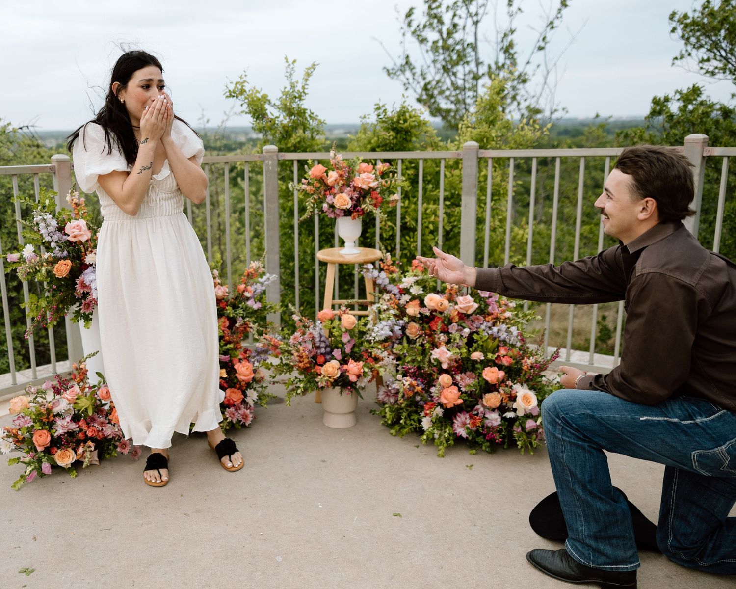 Man proposes on one knee to surprised woman in white dress surrounded by colorful floral arrangements on scenic overlook.