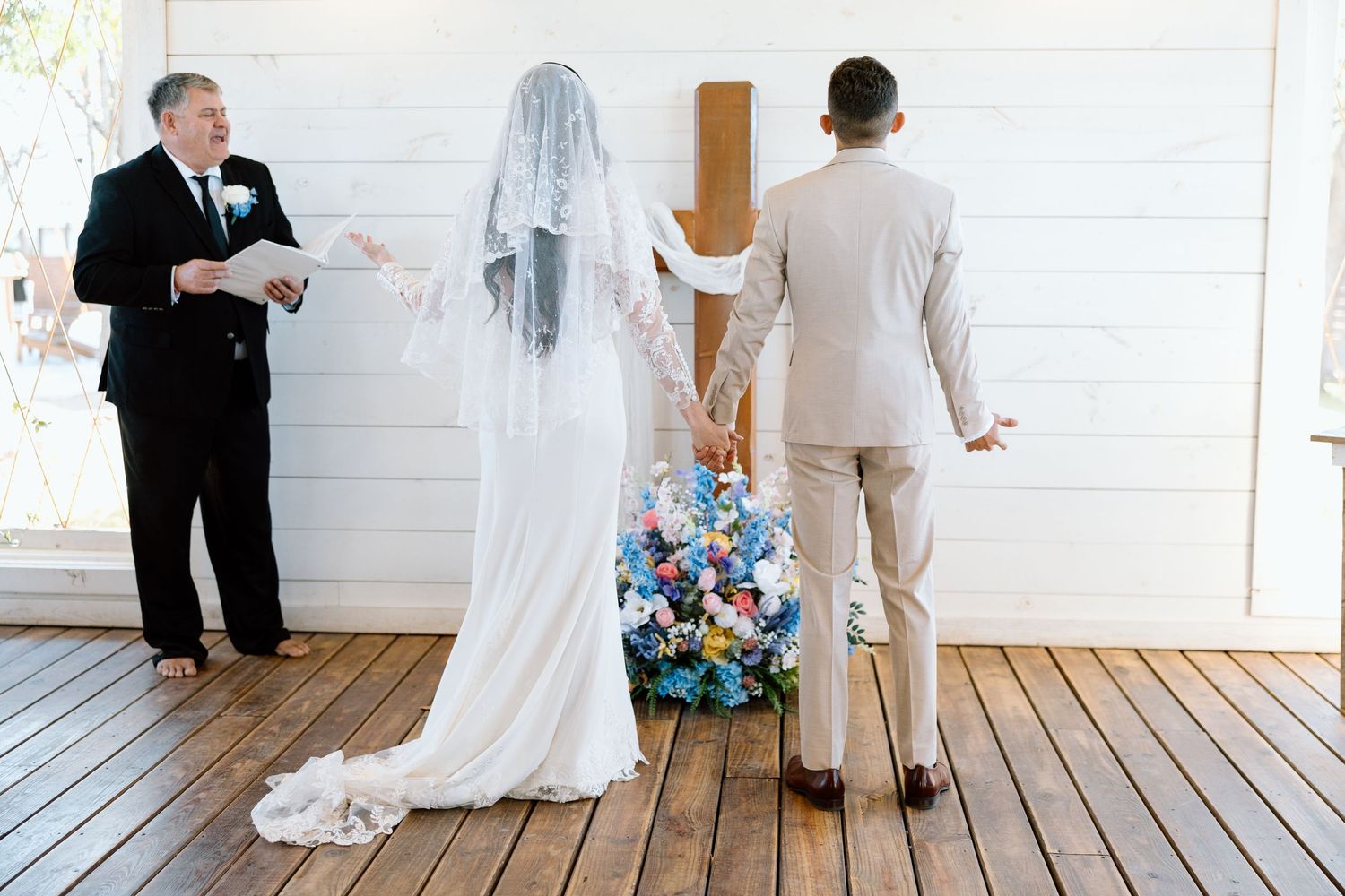 Bride and groom holding hands before wooden cross during outdoor wedding ceremony.