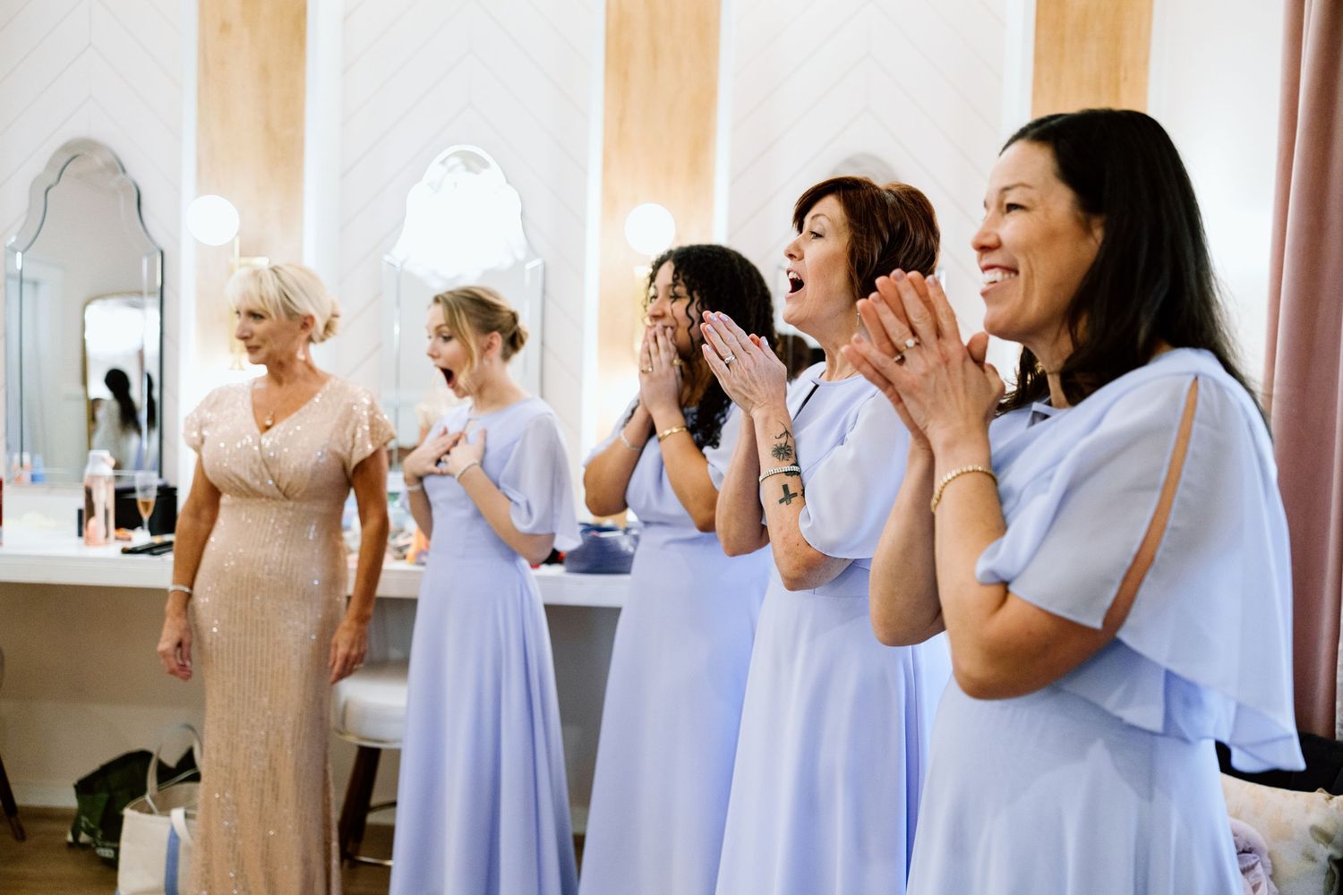 Bridesmaids in lavender dresses react with joy and applause in a bridal suite during wedding preparations.
