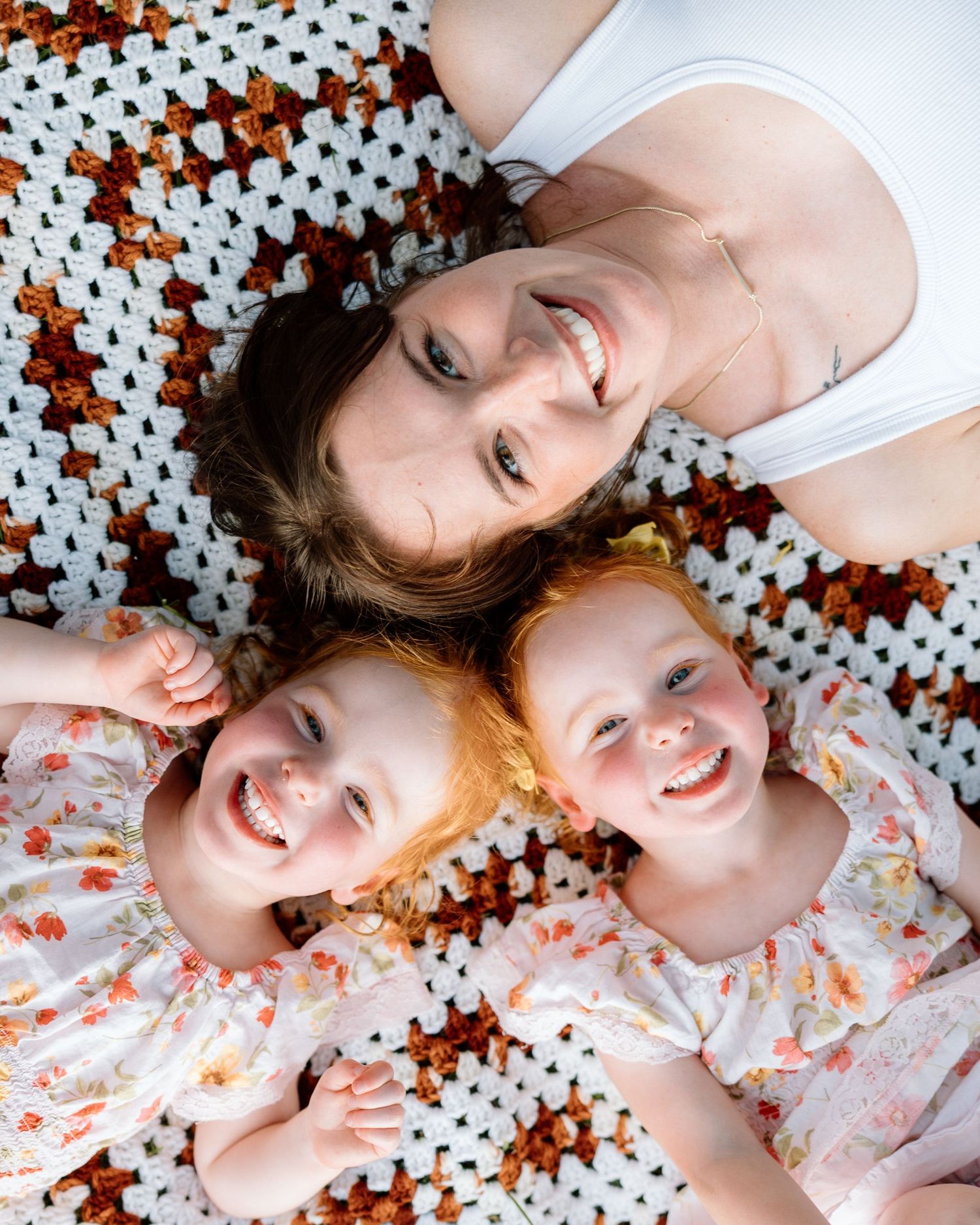A smiling mother and two red-haired toddler girls lying on a crocheted blanket, viewed from above.