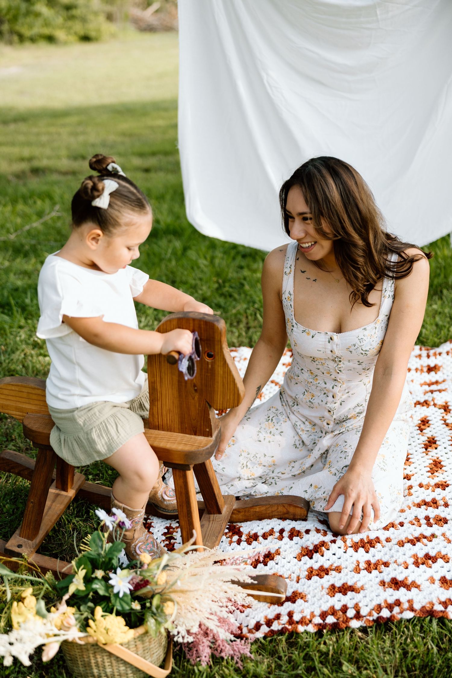 A smiling mother in a floral dress sits on a blanket outdoors with her young daughter playing on a wooden rocking horse.
