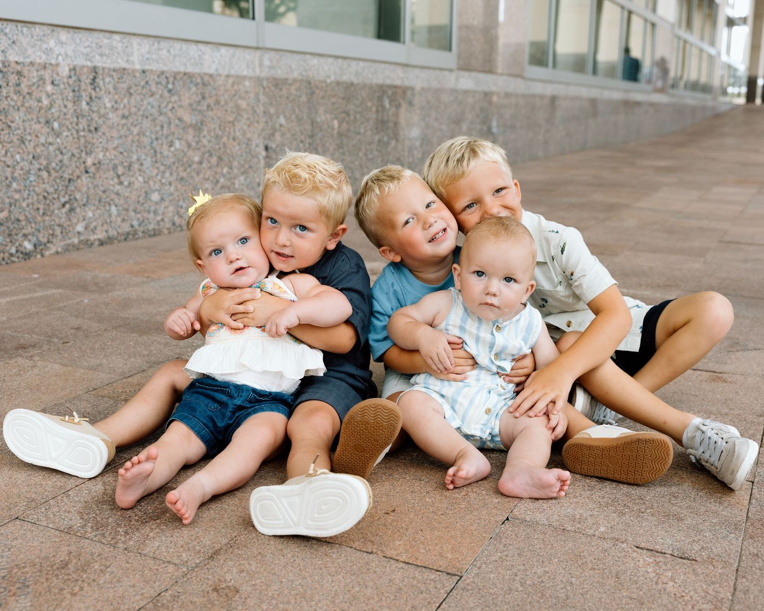 Five young children sit together on a brick sidewalk, smiling and hugging each other in a sweet group photo.