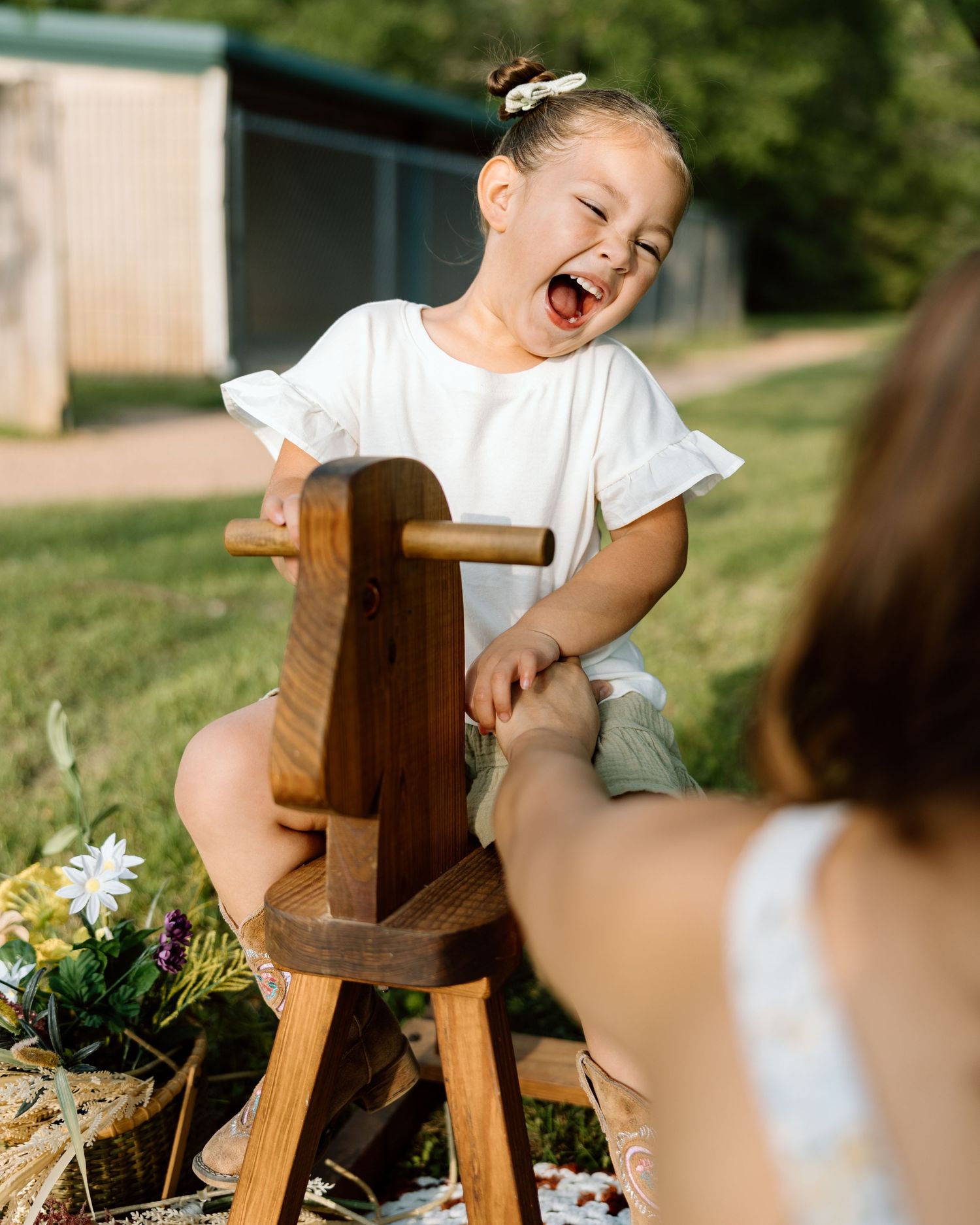 A joyful toddler girl laughs while sitting on a wooden rocking horse outdoors on a sunny day.