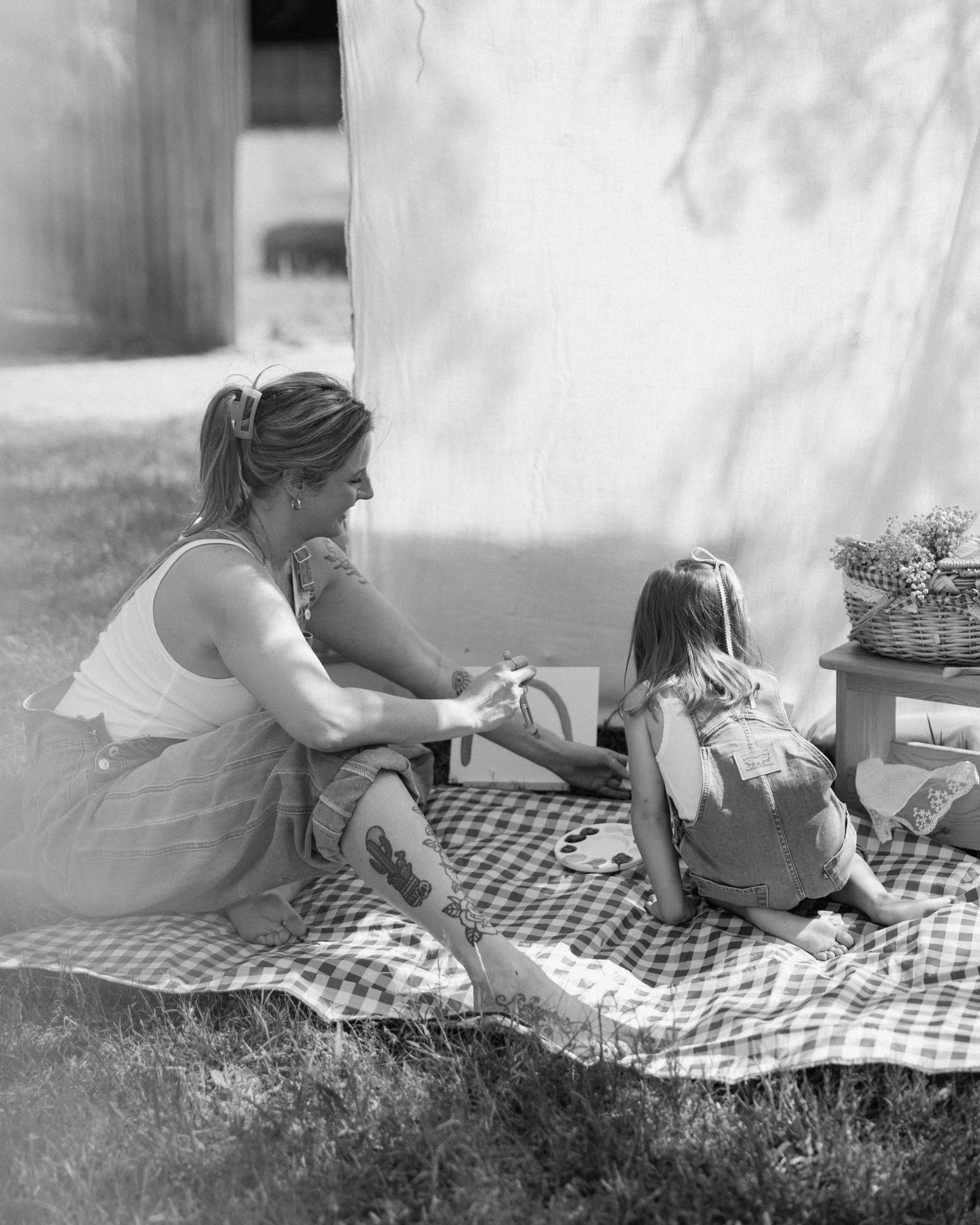 Black and white photo of a woman and young girl sitting on a plaid blanket outdoors, sharing a playful moment together.