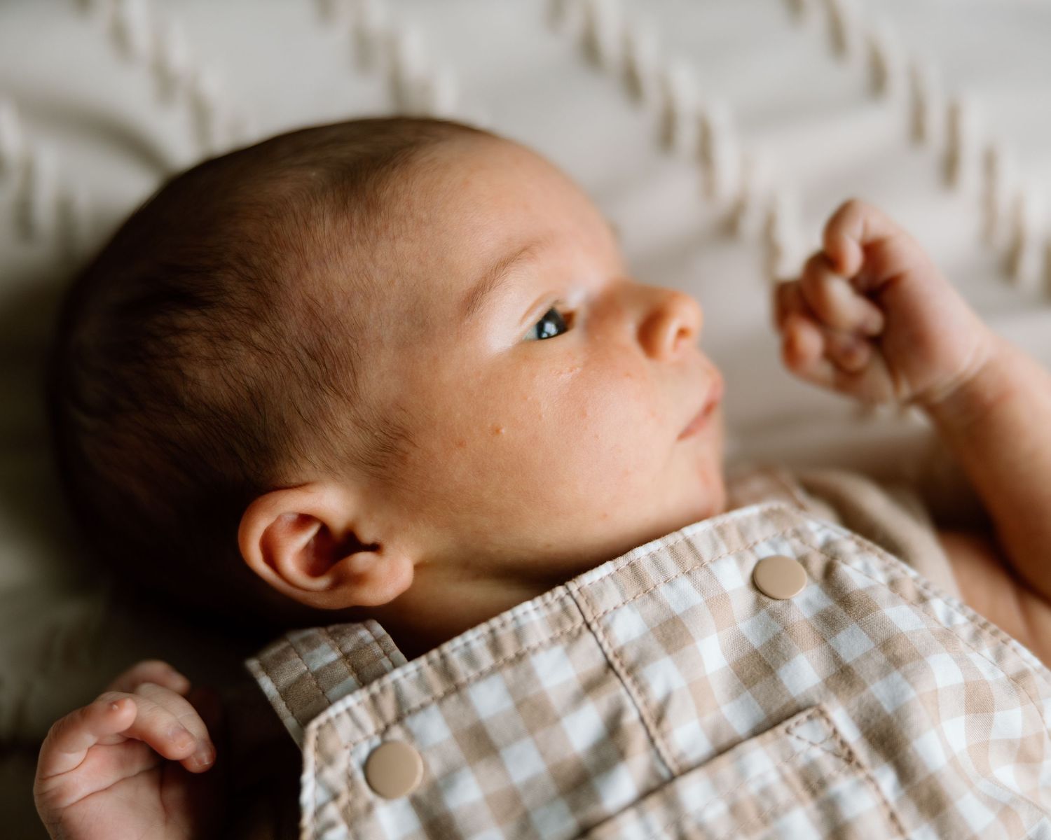 Newborn baby lying on white textured blanket wearing plaid romper, gazing upward with tiny fist raised.