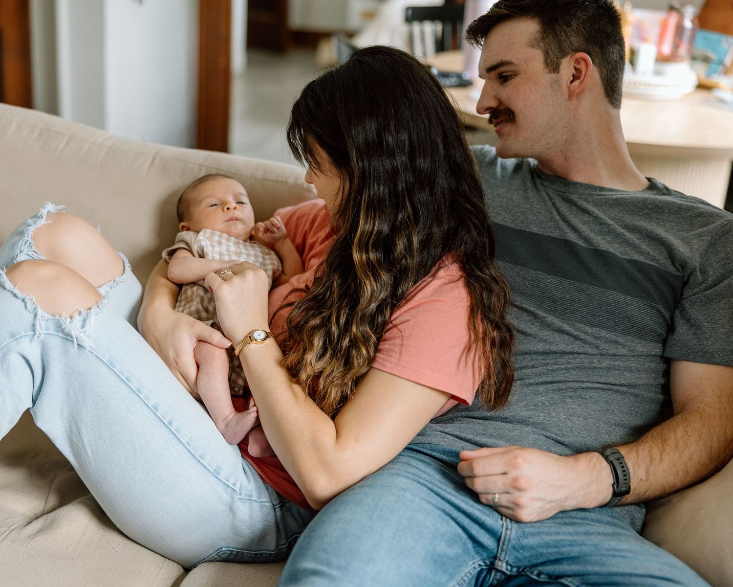 Young couple sitting on couch at home, lovingly looking at their newborn baby held by the mother.