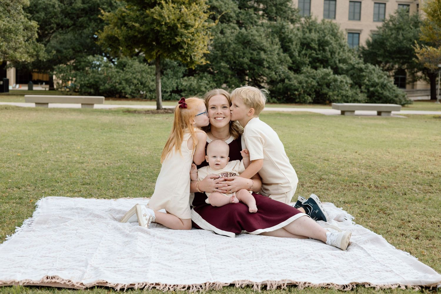 A family sits on a white blanket outdoors, two children kissing their smiling mother on the cheeks while she holds a baby.
