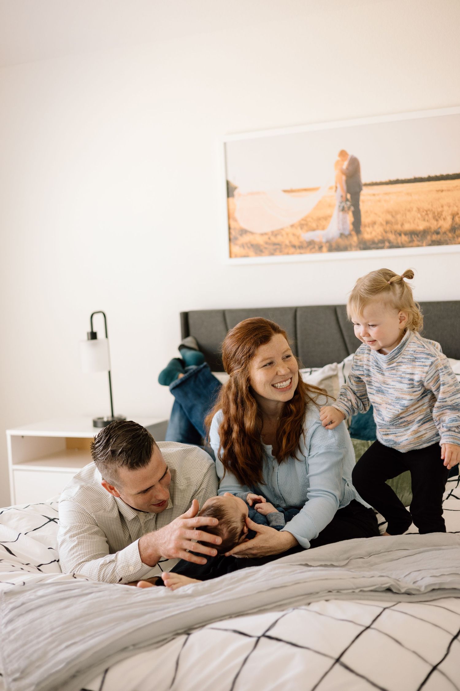 Happy family with toddler and newborn baby bonding on bed in bright modern bedroom with wedding photo on wall.
