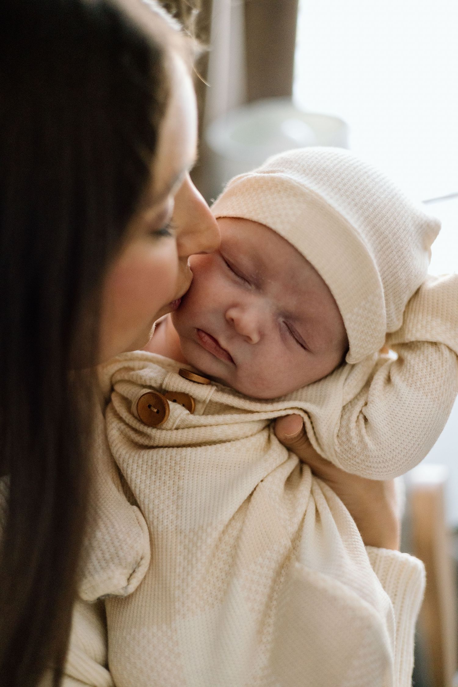 A mother tenderly holds her sleeping newborn baby dressed in a cream knit outfit and matching hat.