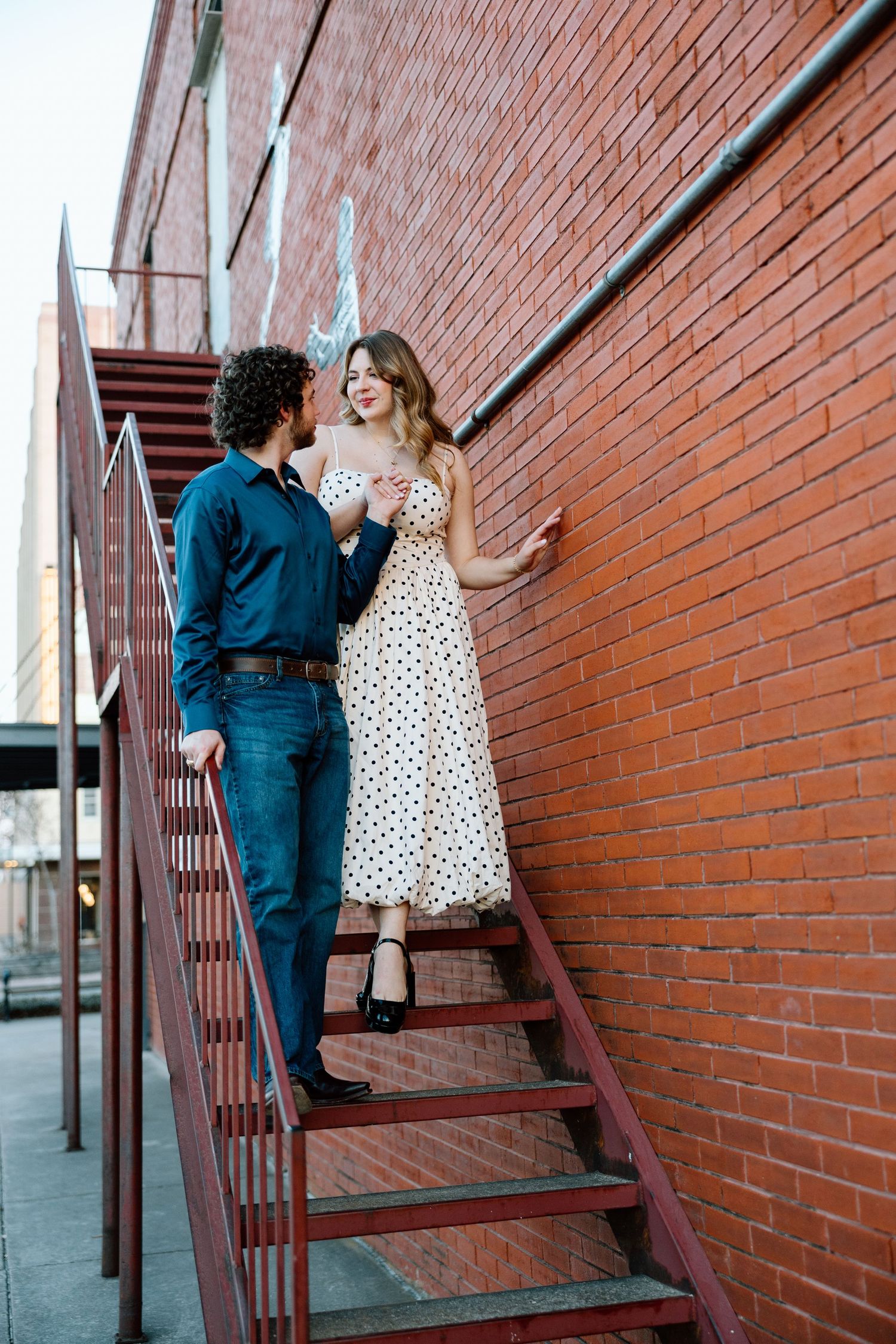 Couple in romantic pose on red metal fire escape stairs against brick wall, woman in polka dot dress.