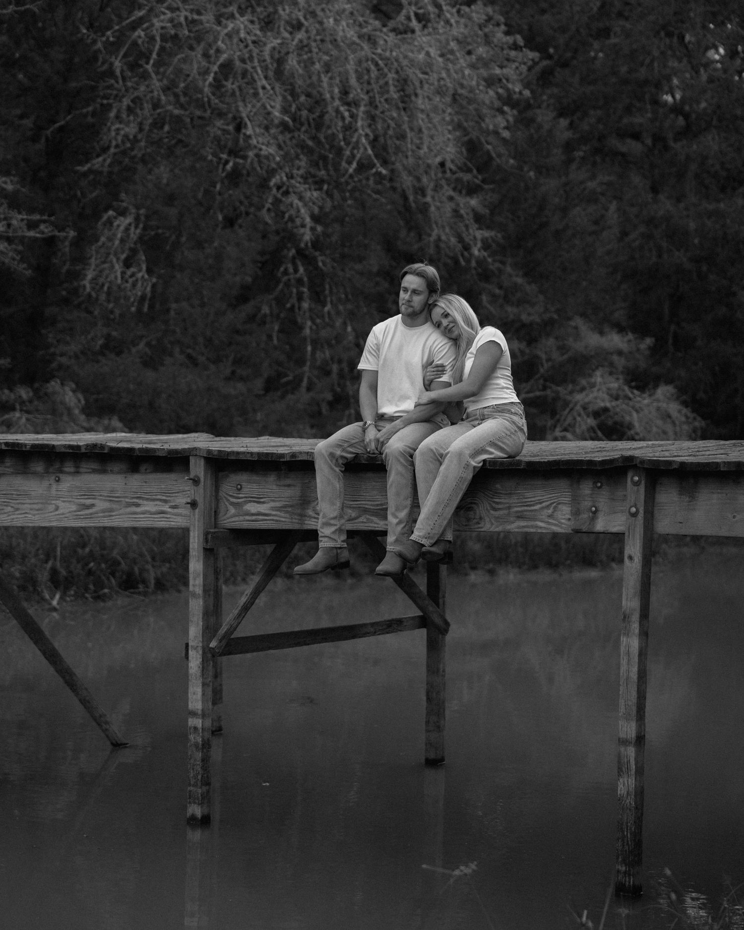 Couple sitting close together on a wooden dock over calm water, surrounded by trees, black and white photo.