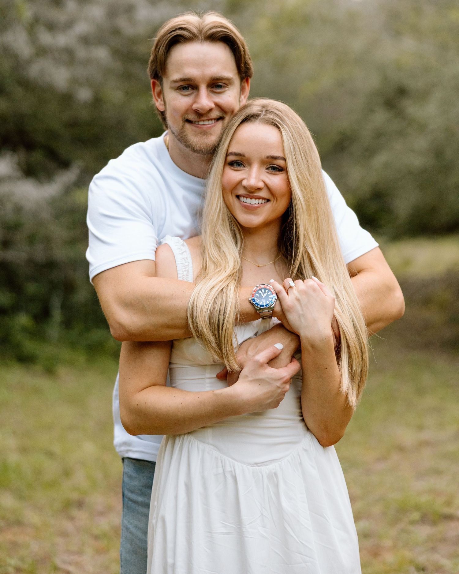 A smiling couple embraces outdoors, woman in white dress showing a ring, man in white tee, surrounded by greenery.