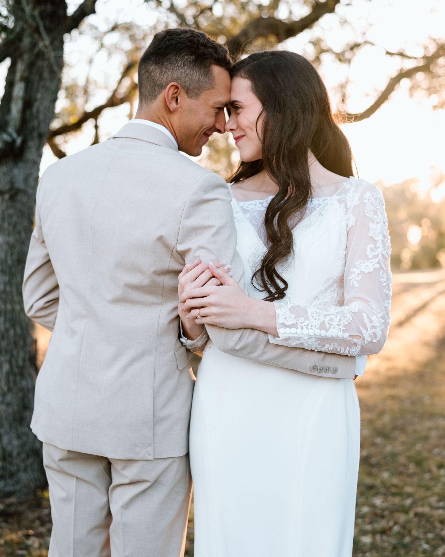 Bride and groom touch foreheads tenderly outdoors at golden hour, bride in lace sleeve wedding gown.