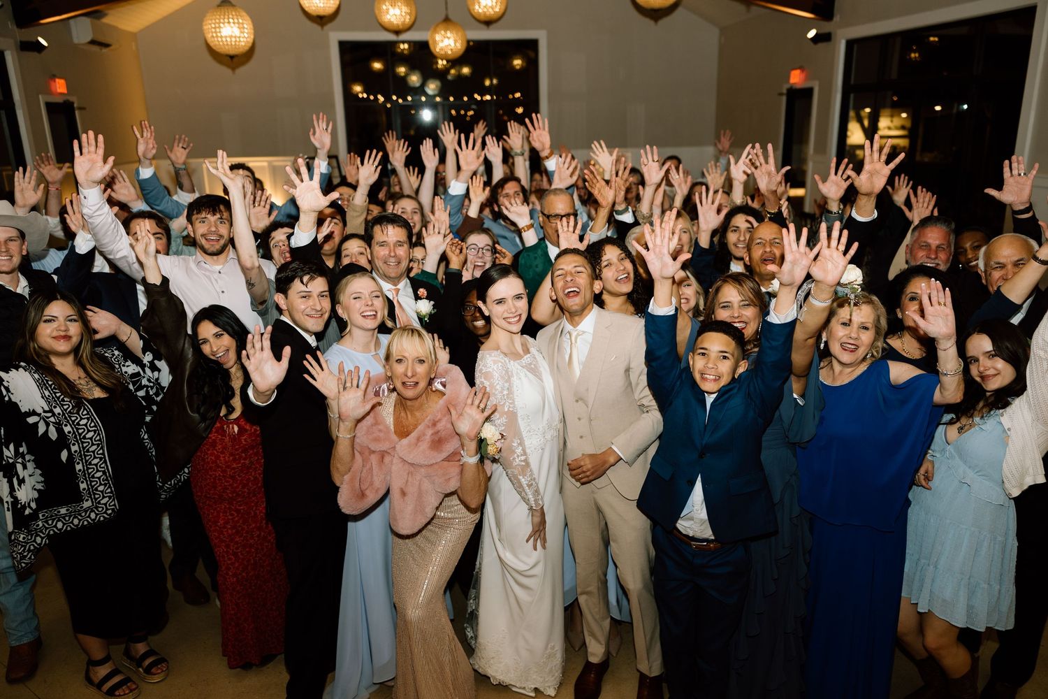 Large group of wedding guests celebrating with hands raised at an indoor wedding reception.