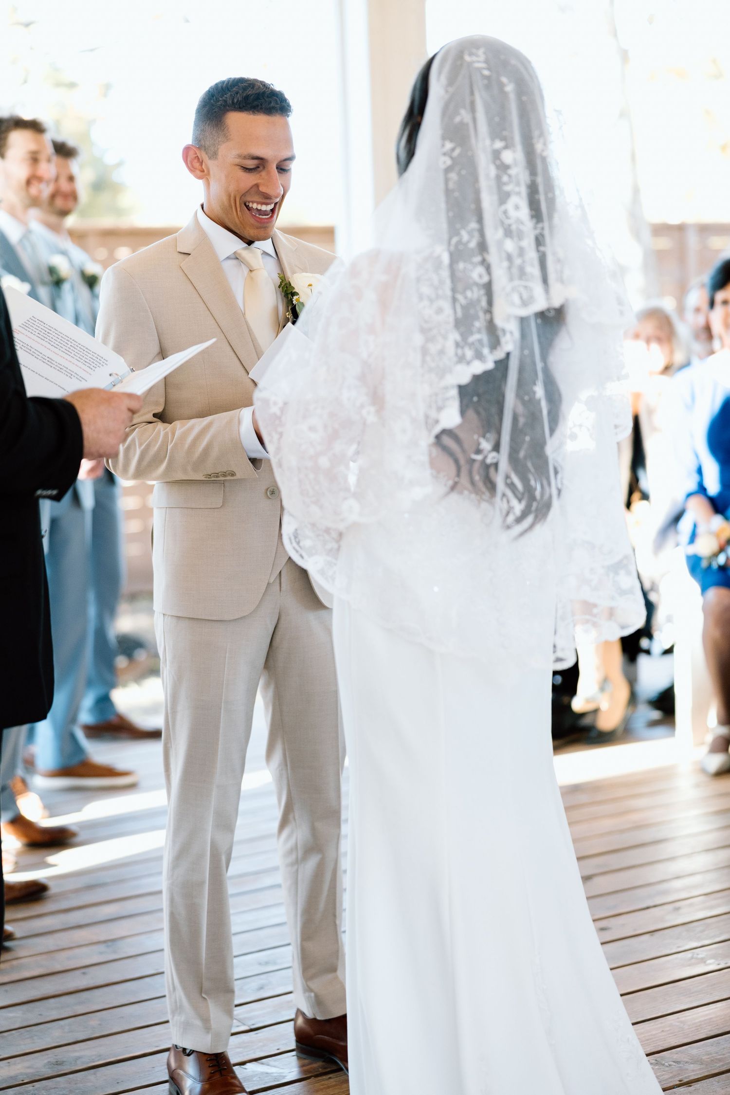 Smiling groom in tan suit reading vows to bride in lace veil at wedding.