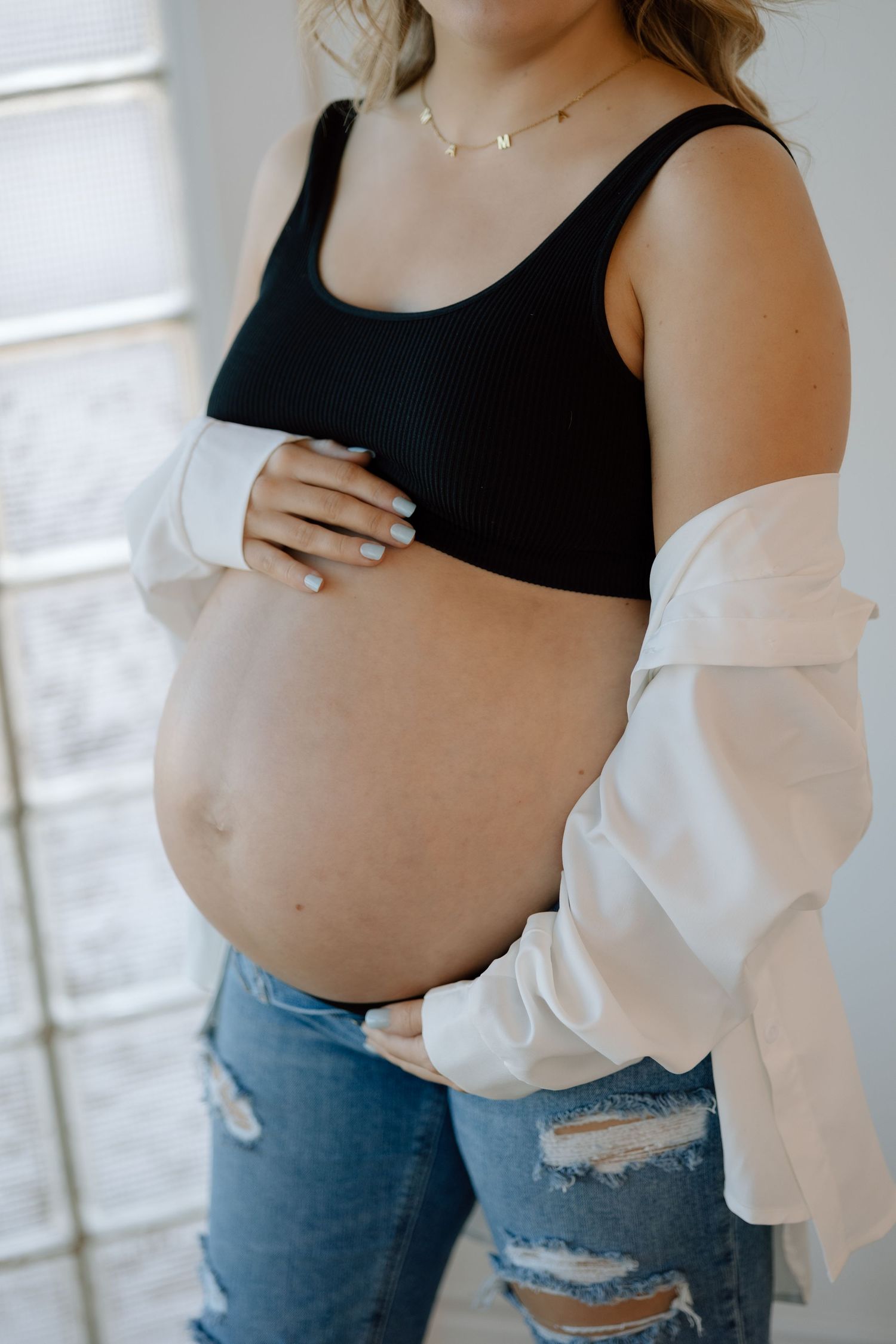 A pregnant woman wearing a black crop top and ripped jeans stands against a white wall in soft lighting.