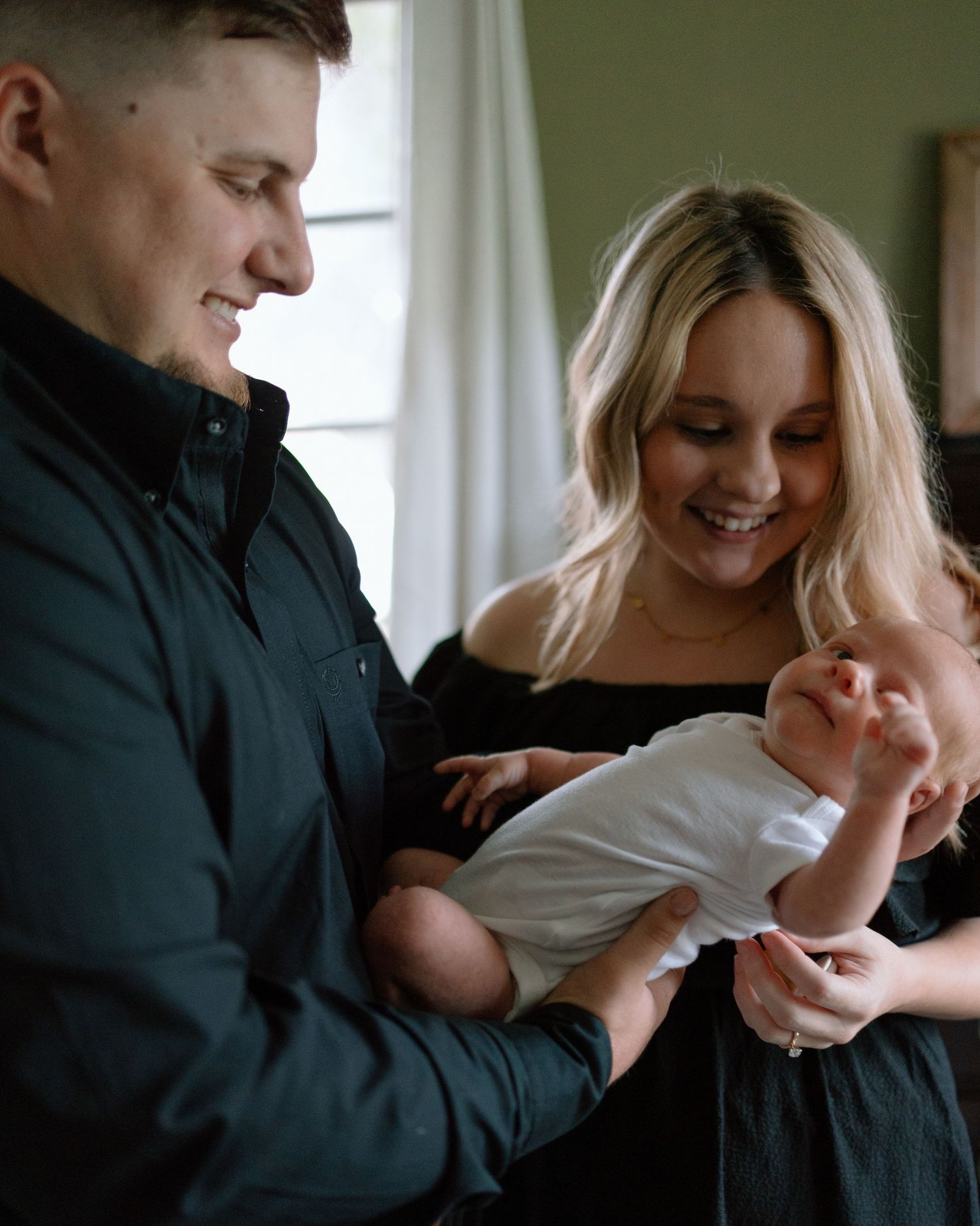 A couple smiles joyfully while holding a small baby in their arms near a window with natural lighting.