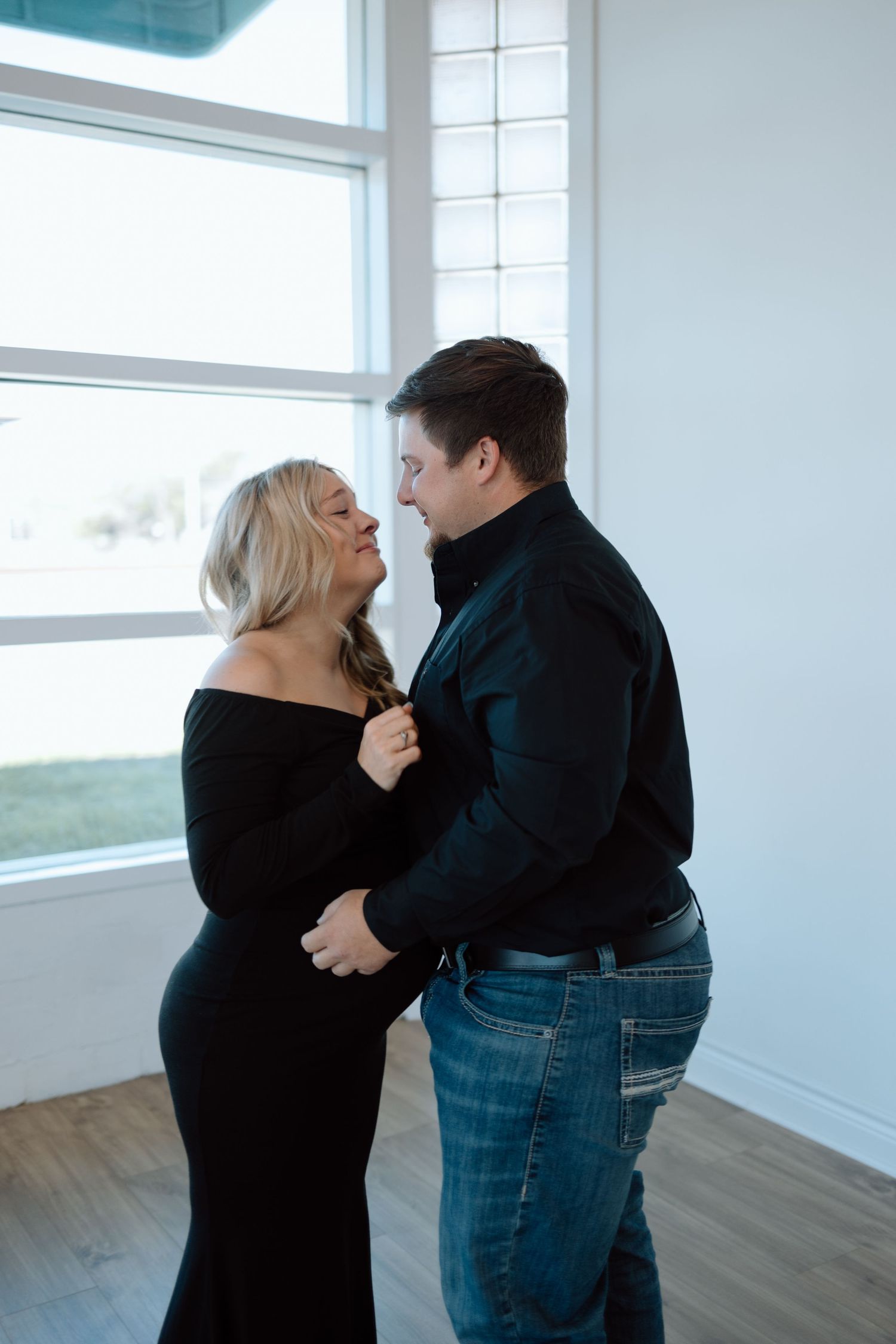 A romantic couple sharing an intimate moment by a window in a bright room, dressed in black clothing.