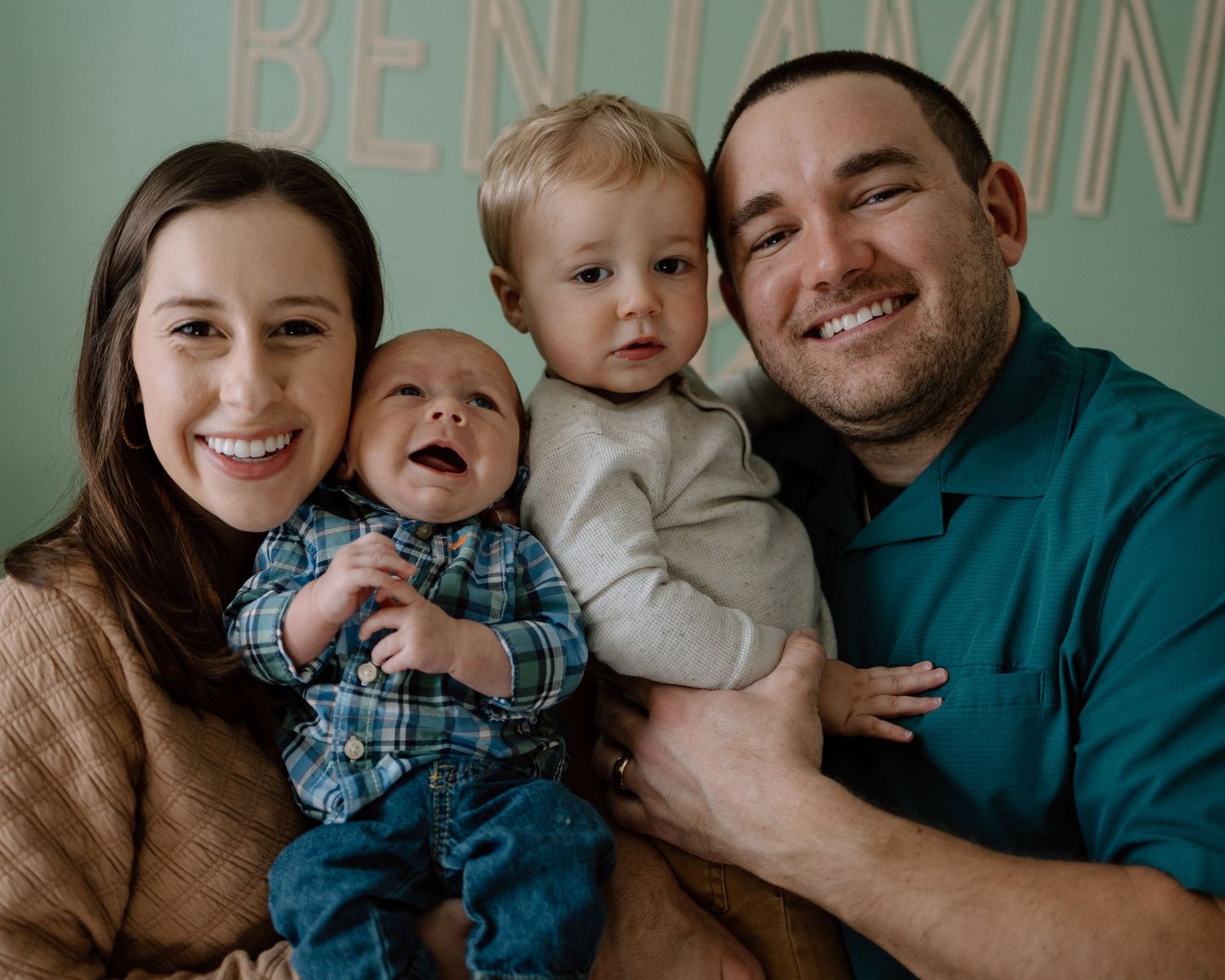 A happy family of four poses together against a mint green wall while smiling warmly at the camera.
