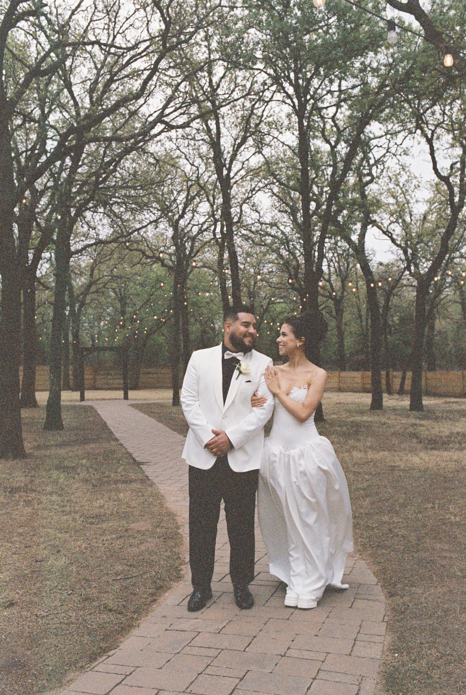 A joyful wedding couple walks together on a brick path through a serene wooded area.