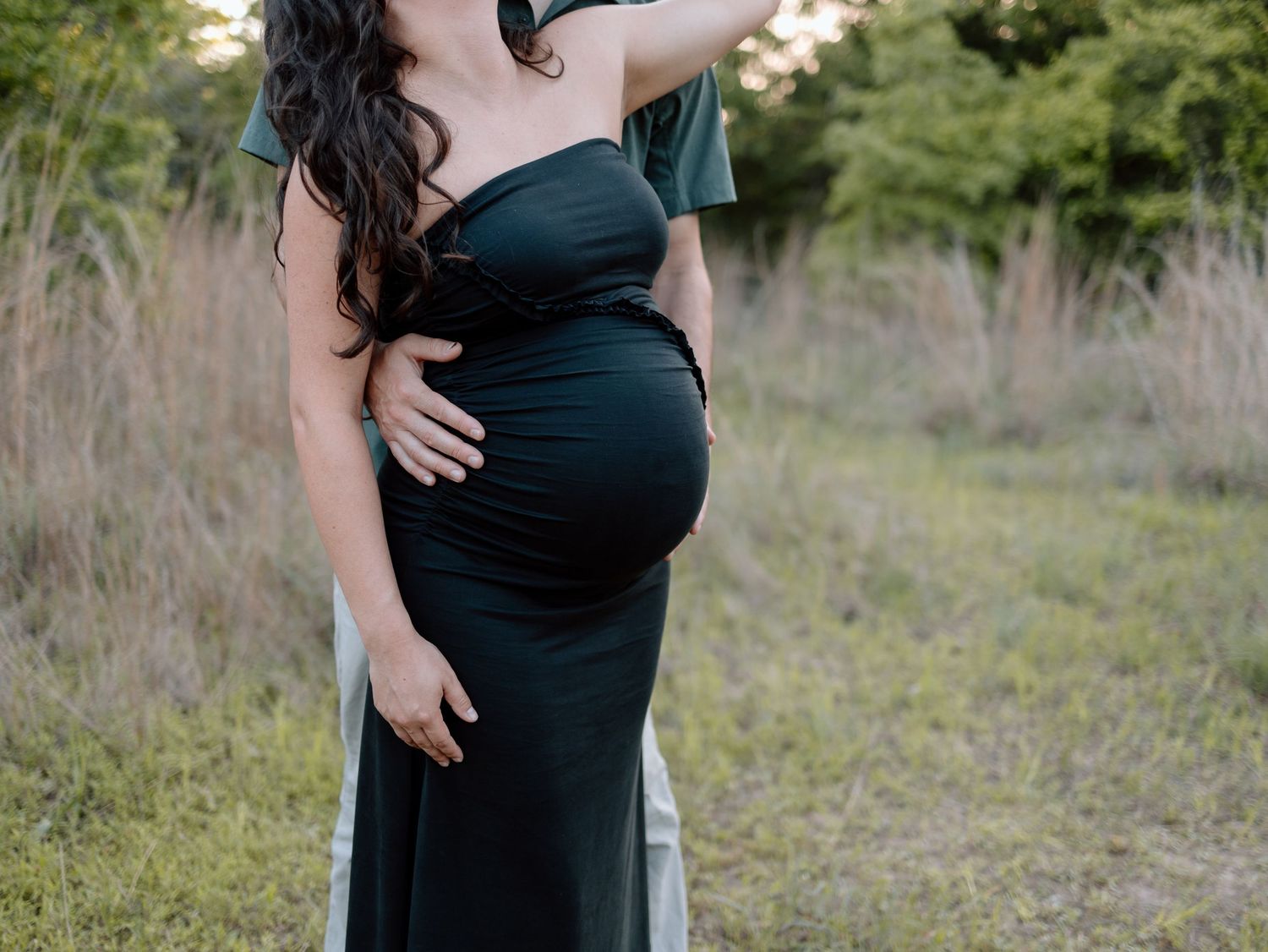 Side view of expecting couple embracing in a field, showing off baby bump in black dress.