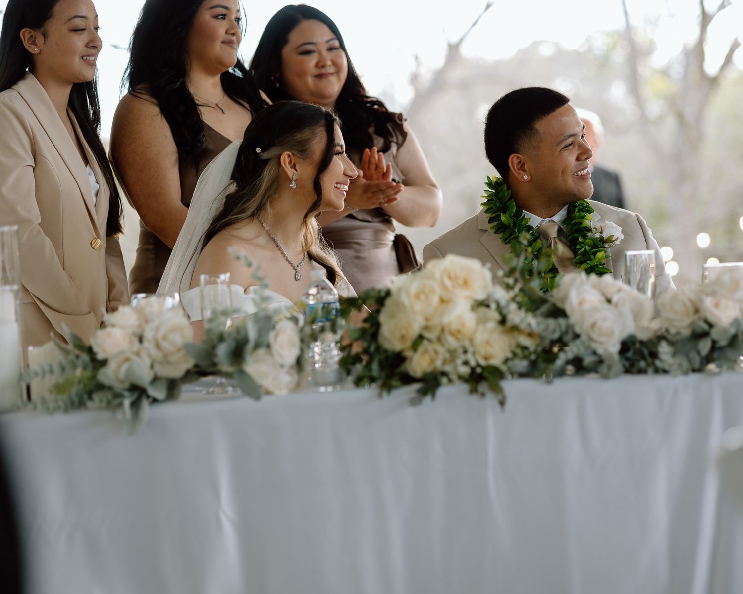 Wedding party seated at decorated head table with white floral arrangements.