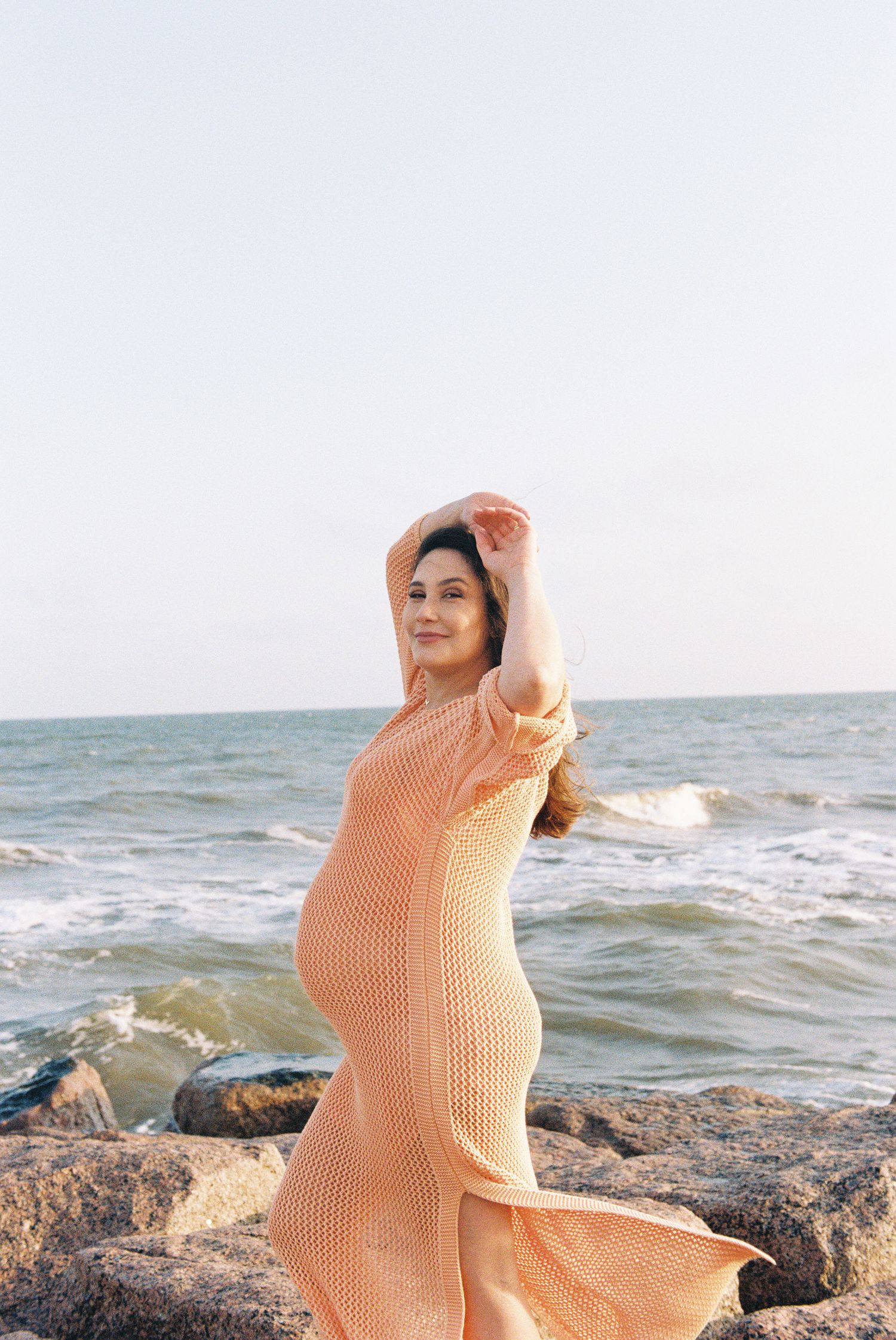 Pregnant woman in peachy dress poses gracefully on beach rocks.
