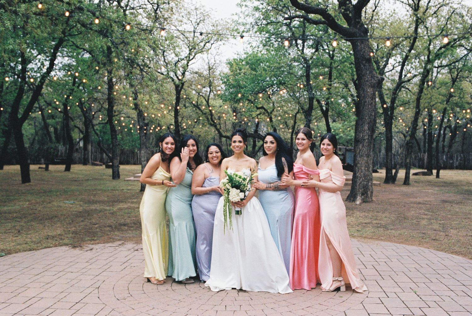 Wedding party poses together outdoors wearing pastel colored dresses in a wooded setting.