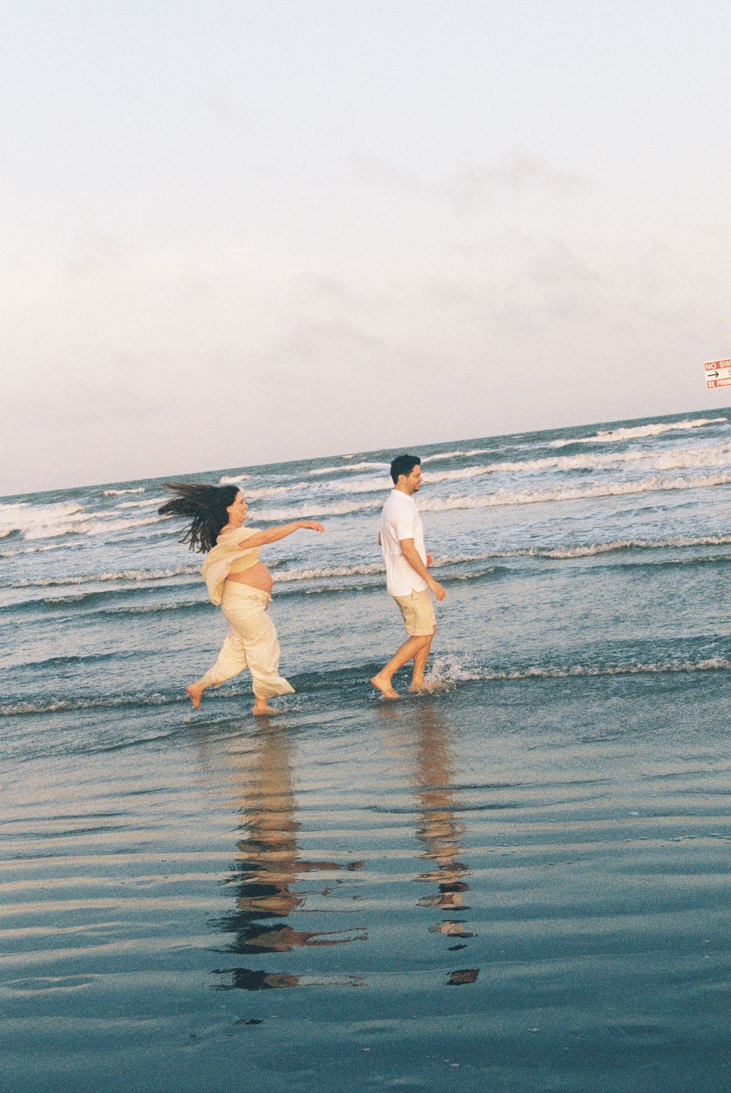 Couple walking along shoreline at sunset during beach maternity photo session.