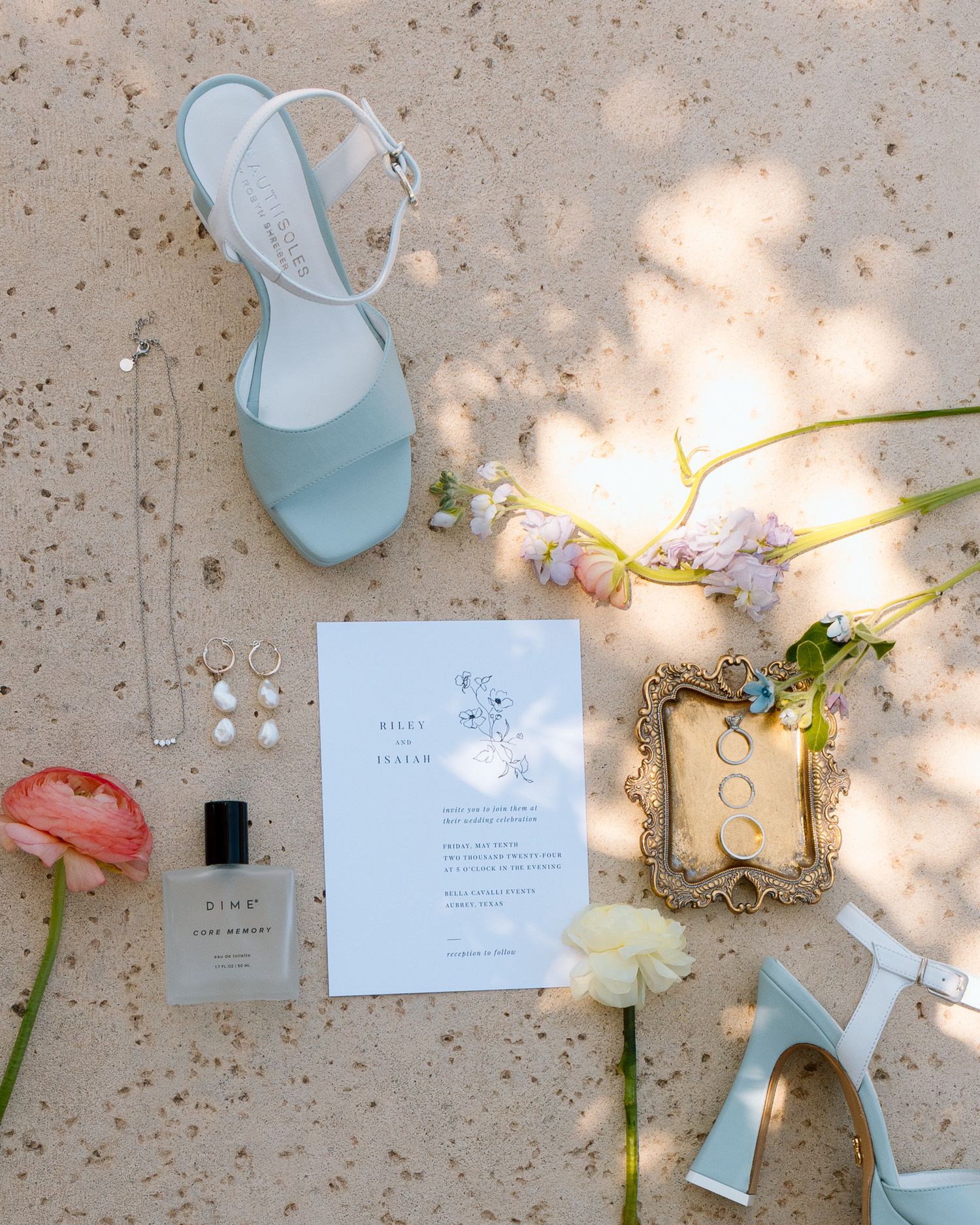 Wedding details flat lay with blue shoes, invitation and scattered flowers on textured surface.