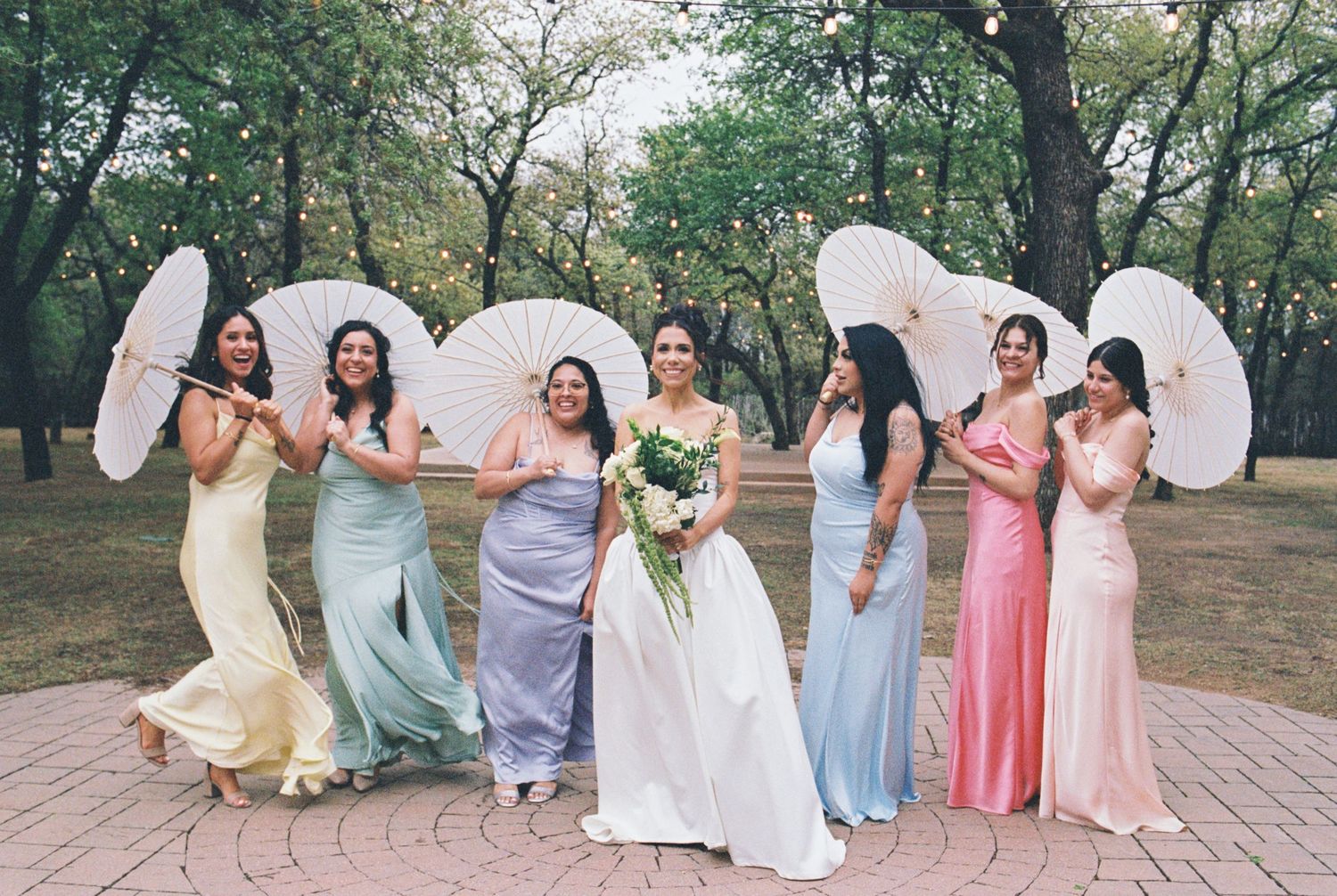 Wedding party holding parasols poses outdoors in pastel dresses on pink stone path.