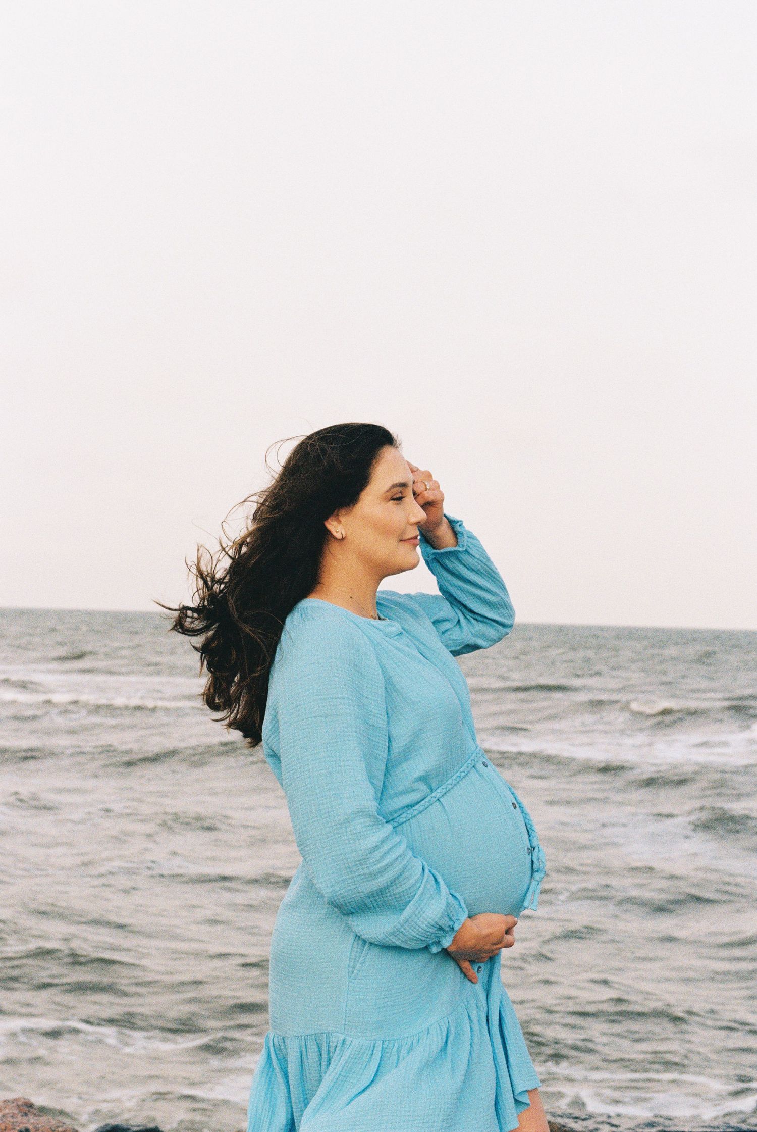 Pregnant woman in blue dress stands by ocean during maternity shoot.