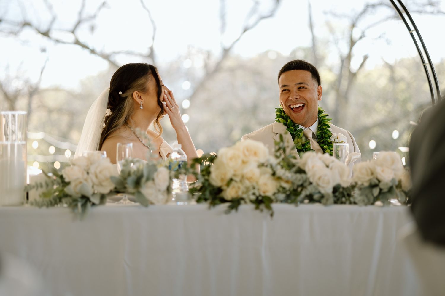 Couple shares joyful moment at white-decorated wedding reception table.