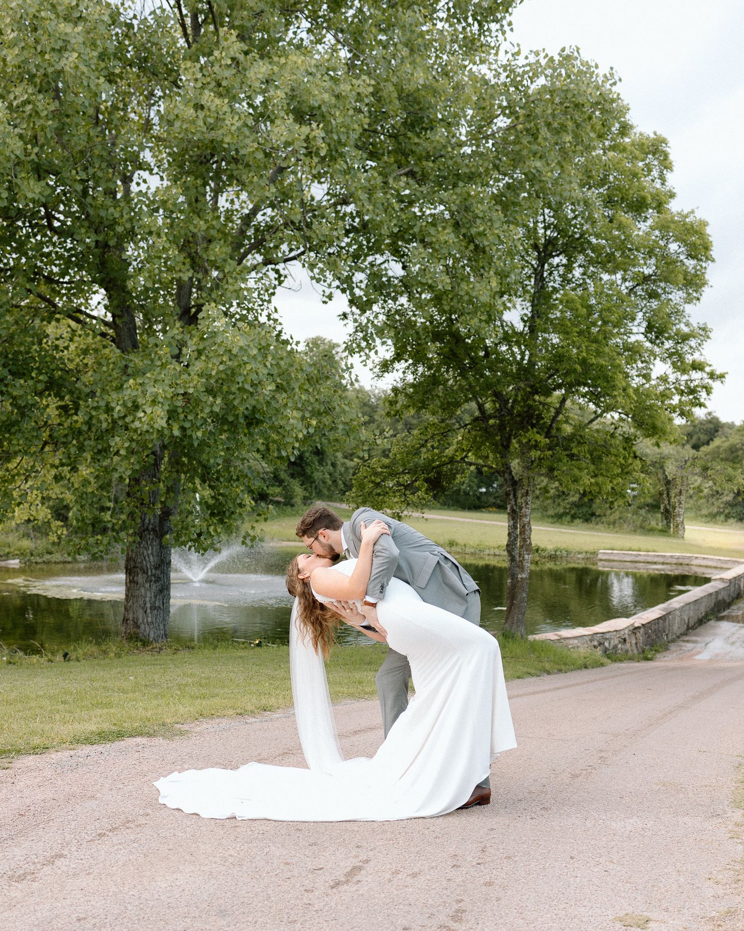 Romantic dip pose of newlyweds on path with fountain and trees in background.