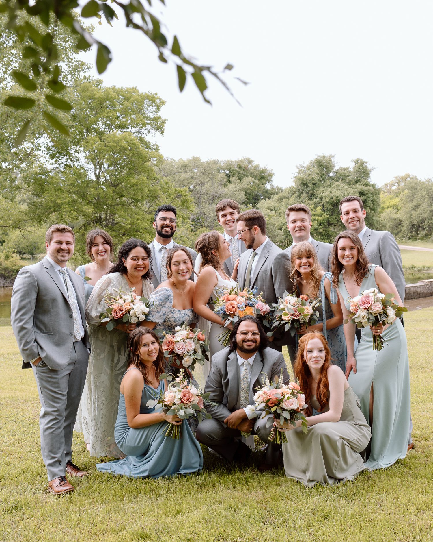 Cheerful wedding party in blue and grey attire gathered for group photo outdoors.
