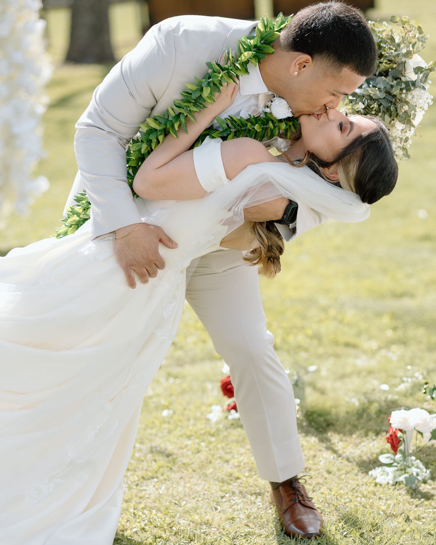 Newlyweds share a passionate dip kiss on their wedding day while wearing traditional Hawaiian green lei garlands.