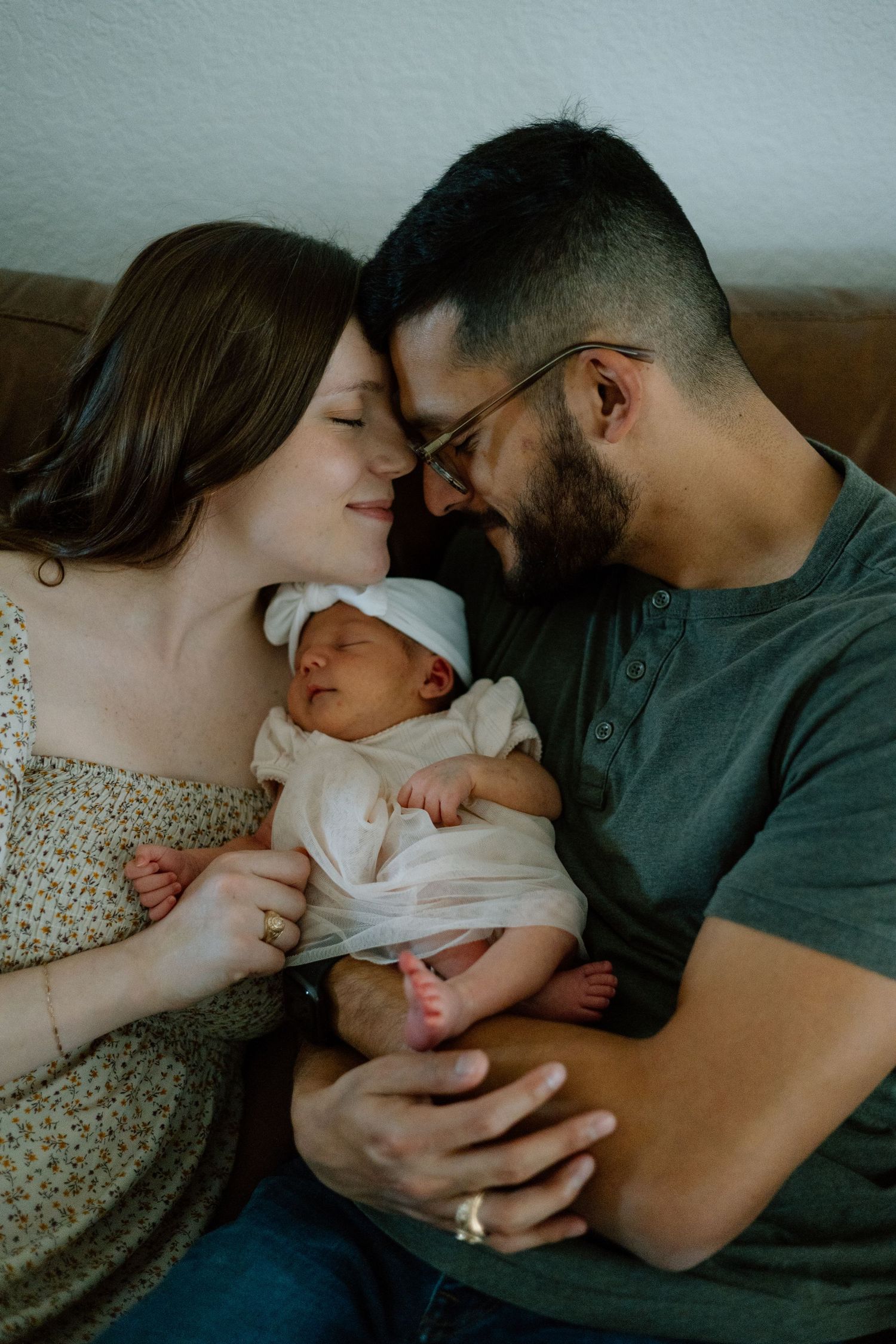 Parents snuggle close together on couch while cradling their sleeping newborn.