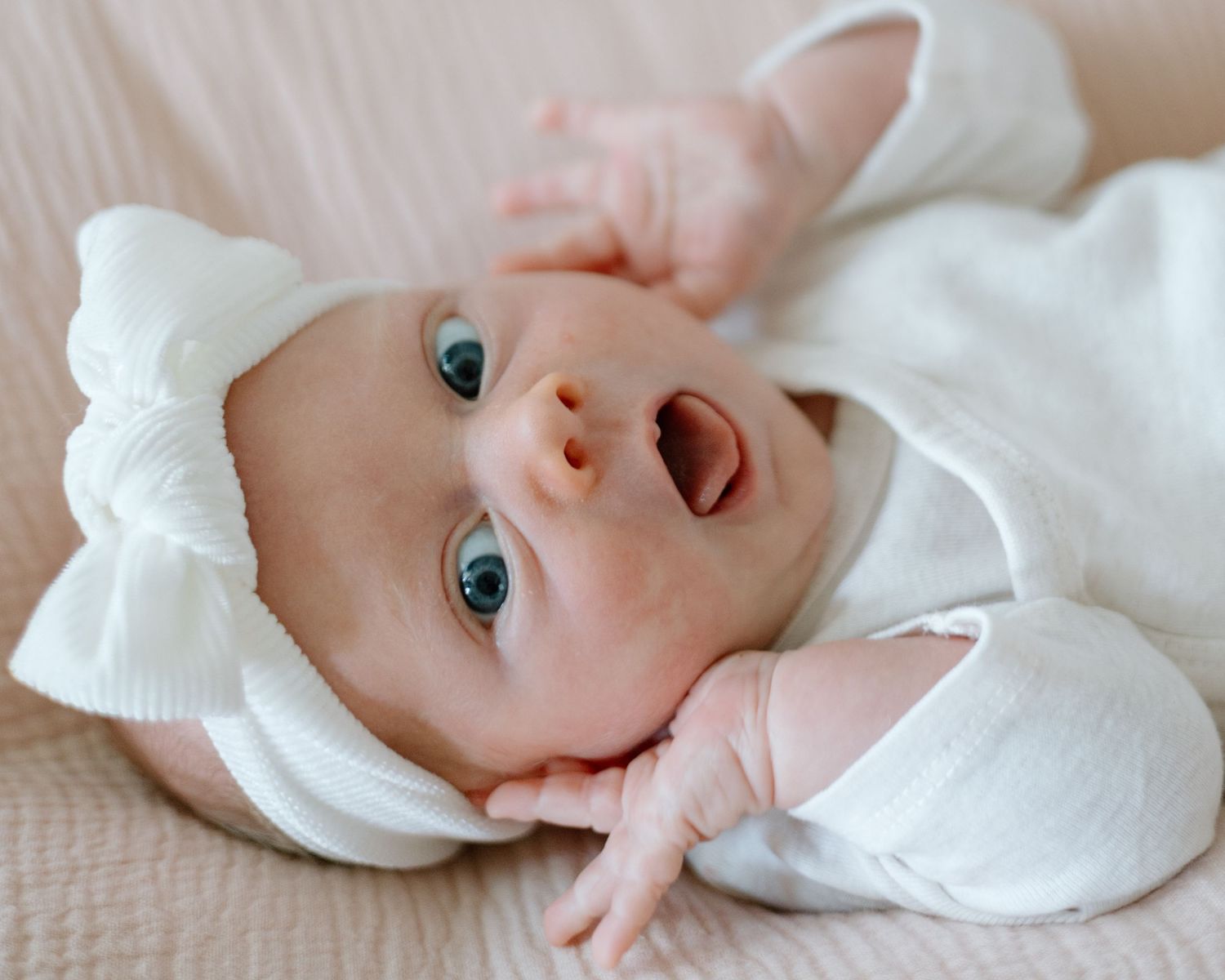 Smiling baby with bright blue eyes laying on white blanket looking up at camera.